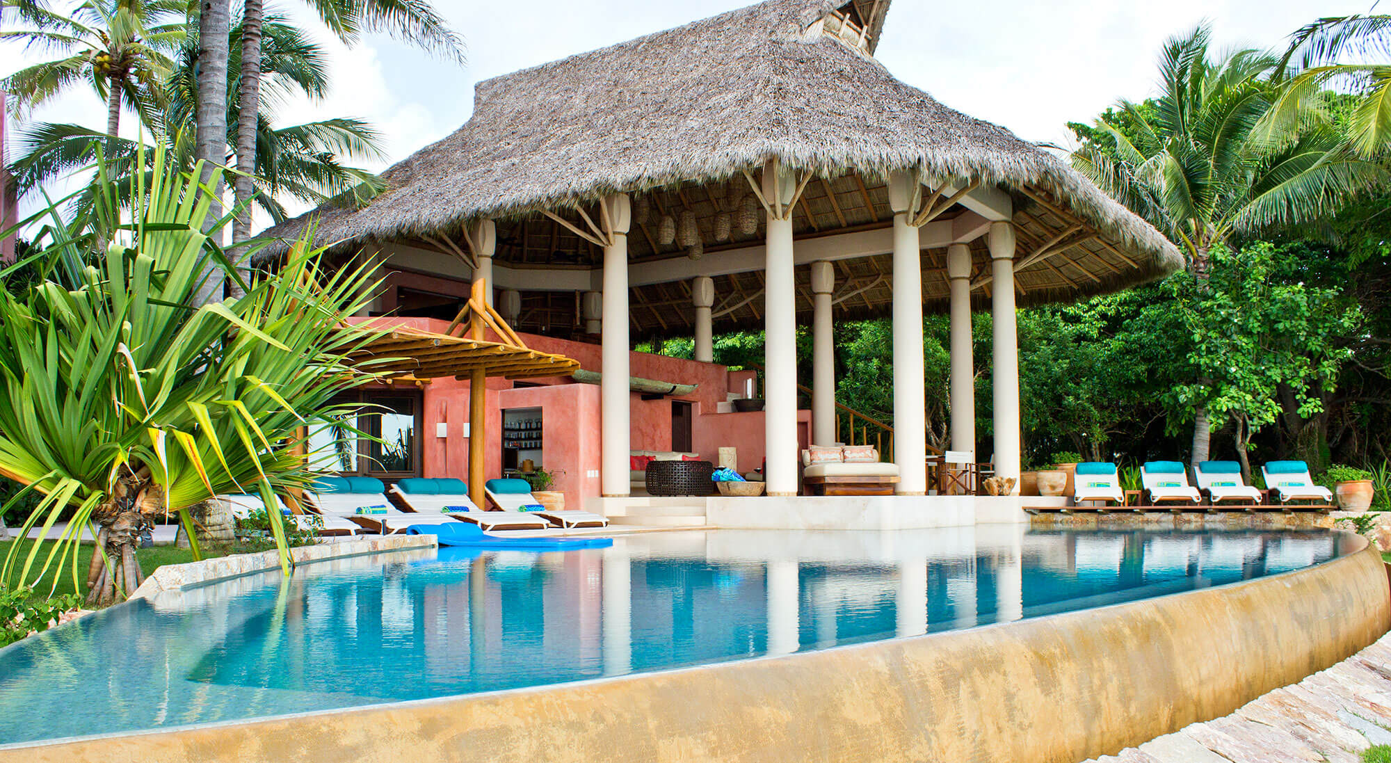 Daytime exterior of the full estate — the circular thatched palapa pavilion with round stucco columns at center, white teak loungers with teal cushions lining the curved pool on both sides, a bamboo-and-log shade structure at left, the terracotta building with upper balcony rising behind, tall palms and tropical garden surrounding, Estate Venado, Ranchos Estates, Punta Mita.