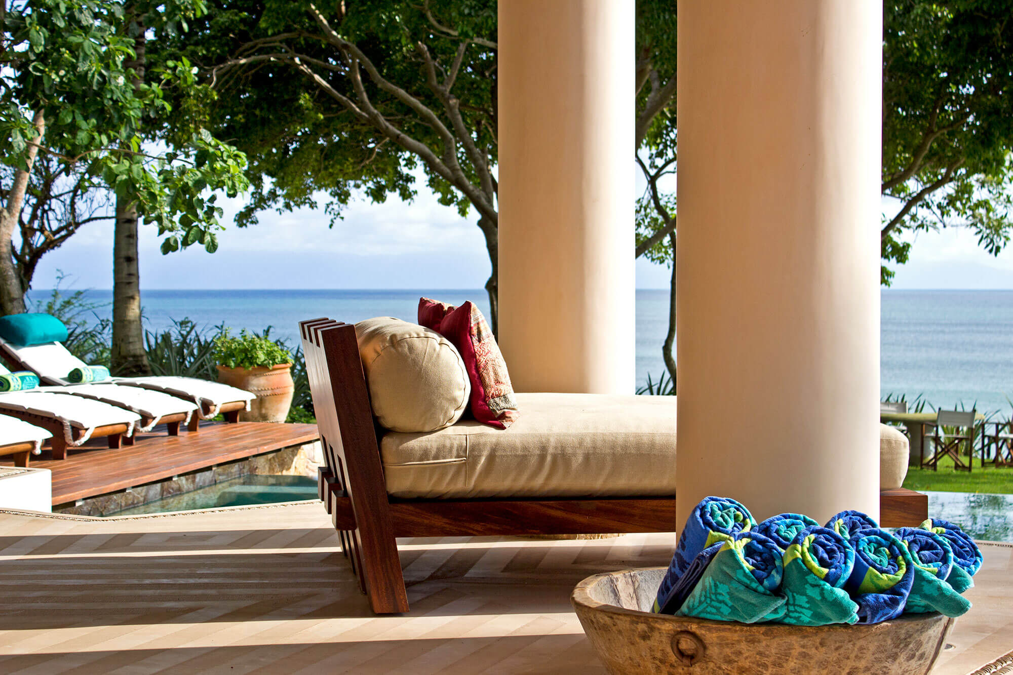 Covered terrace with a teak daybed and cream cushion beside a round stone bowl filled with rolled teal and blue towels, round cream stucco columns framing the view of the pool, garden trees, and open Pacific beyond, Estate Venado, Ranchos Estates, Punta Mita.