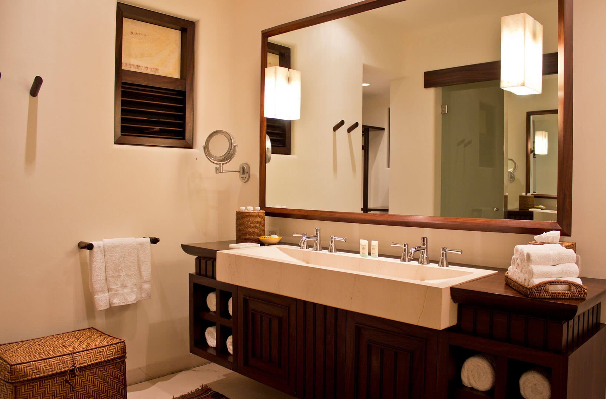 Ensuite bathroom vanity — a wide cream-stone double trough sink set into a dark mahogany cabinet with open towel shelving, a large wood-framed mirror with flanking sconces, an articulated magnifying mirror, dark louvered window, and a woven rattan laundry hamper beside a basket of folded white towels, Estate Venado, Ranchos Estates, Punta Mita.