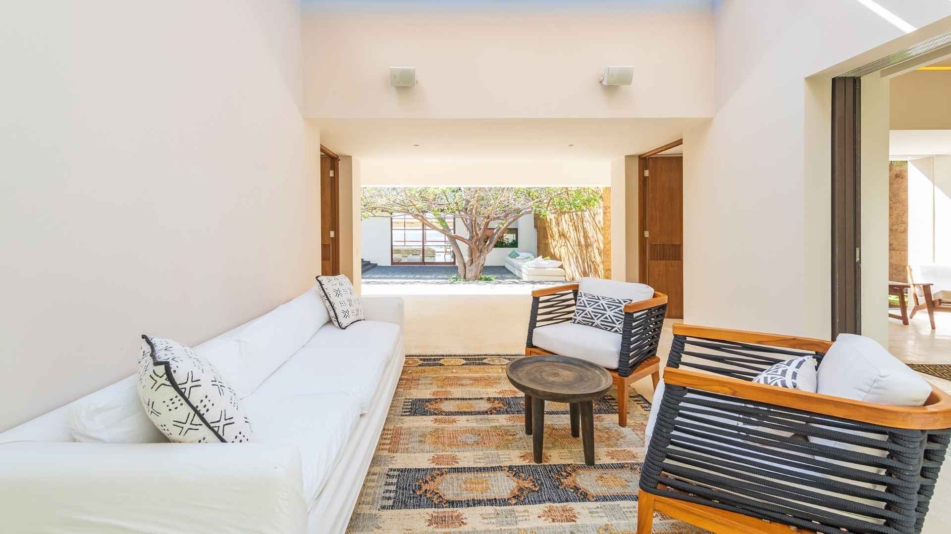 Interior living pavilion from a second angle, showing the white sofa with mudcloth-print pillows, two teak rope chairs, a kilim rug, open archway to the courtyard tree, and a bedroom wing entrance at right, Villa Luna Mar, Ranchos Estates, Punta Mita.