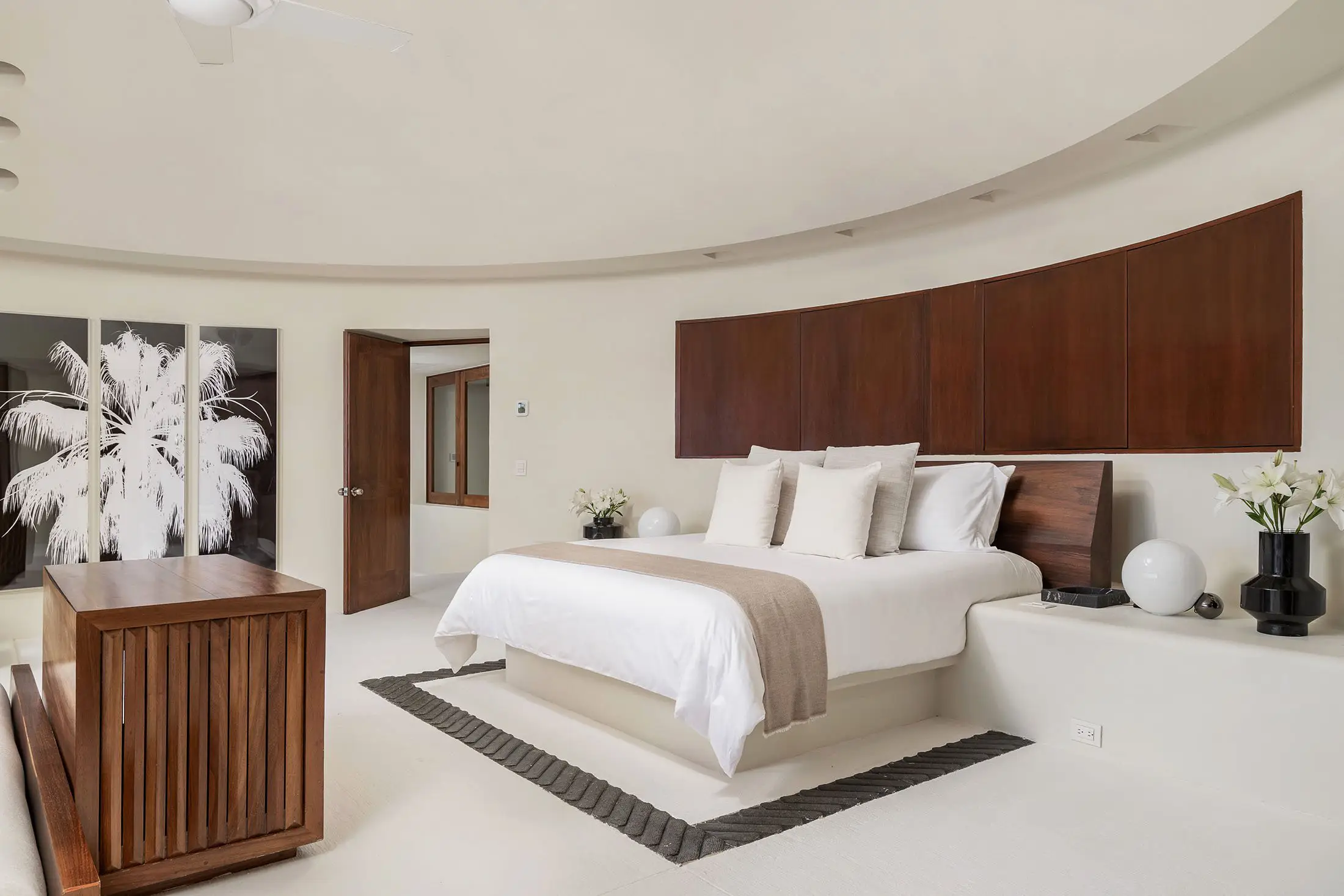 Curved ceiling primary suite with walnut headboard wall, white bedding, and palm-print artwork, Villa Pacífica, Ranchos Estates, Punta Mita.