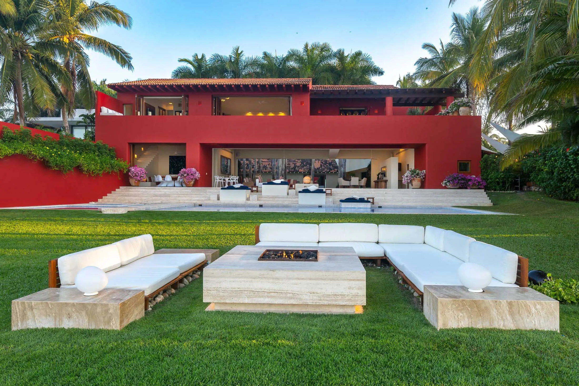 Outdoor fire pit lounge on manicured lawn with red villa facade and terraced stairs beyond, Villa Pacífica, Ranchos Estates, Punta Mita.