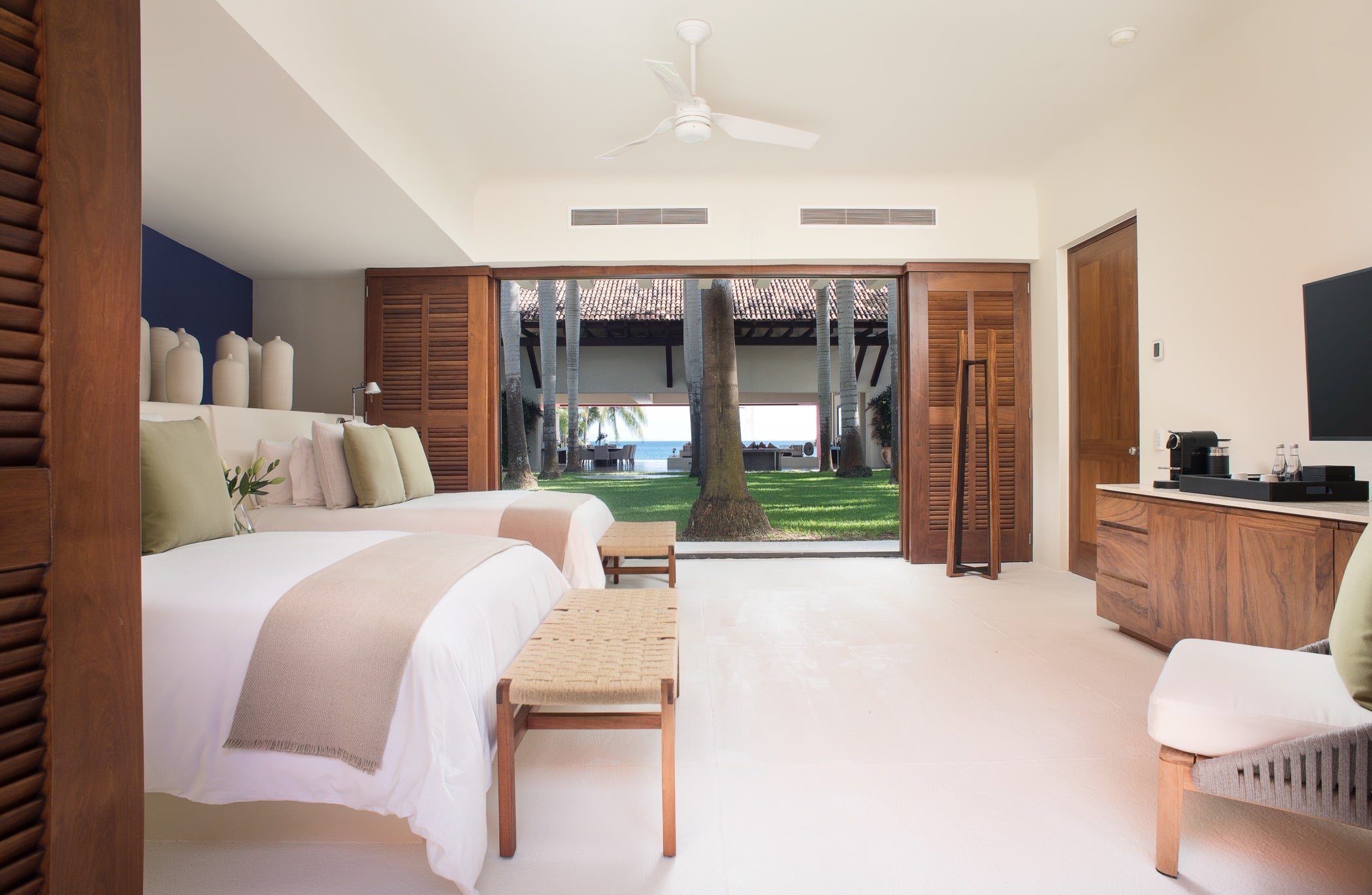 Guest suite with two queen beds, louvered folding doors open to a palm-shaded courtyard and ocean glimpse beyond, Villa Pacífica, Ranchos Estates, Punta Mita.