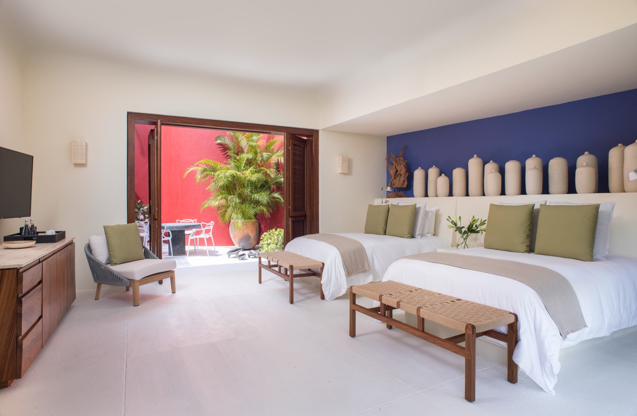 Guest suite with two queen beds, indigo accent wall, and doors open to a red-walled courtyard with potted palms, Villa Pacífica, Ranchos Estates, Punta Mita.