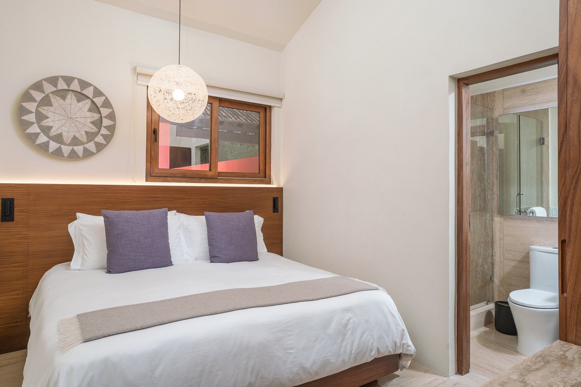 Guest suite king bed with walnut headboard panel and pendant lamp beside an open ensuite with glass shower, Villa Pacífica, Ranchos Estates, Punta Mita.