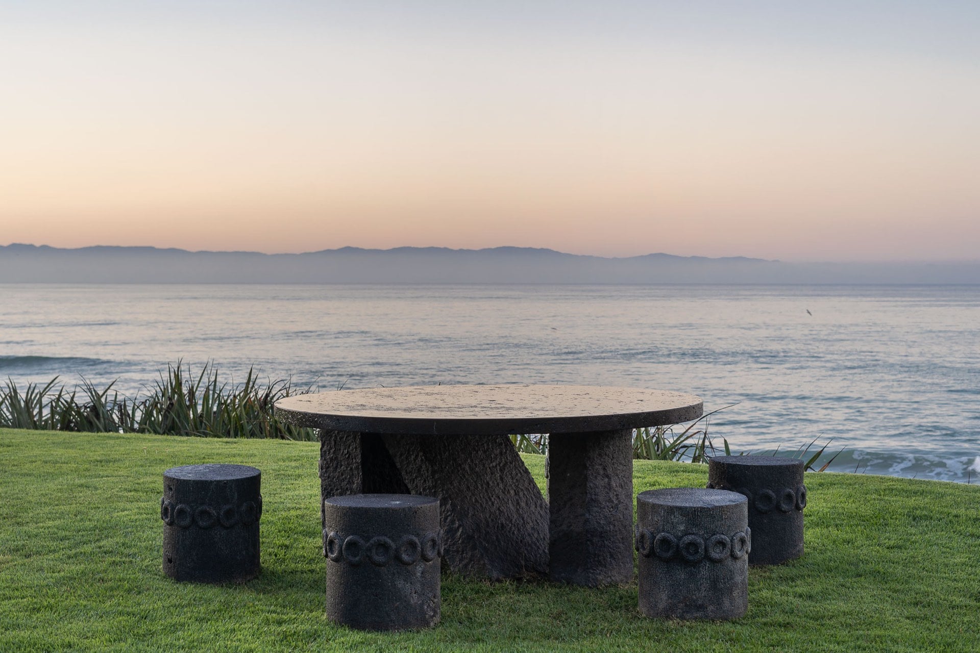 Carved stone outdoor dining table with four cylindrical stools on a beachfront lawn at sunset, Villa Pacífica, Ranchos Estates, Punta Mita.
