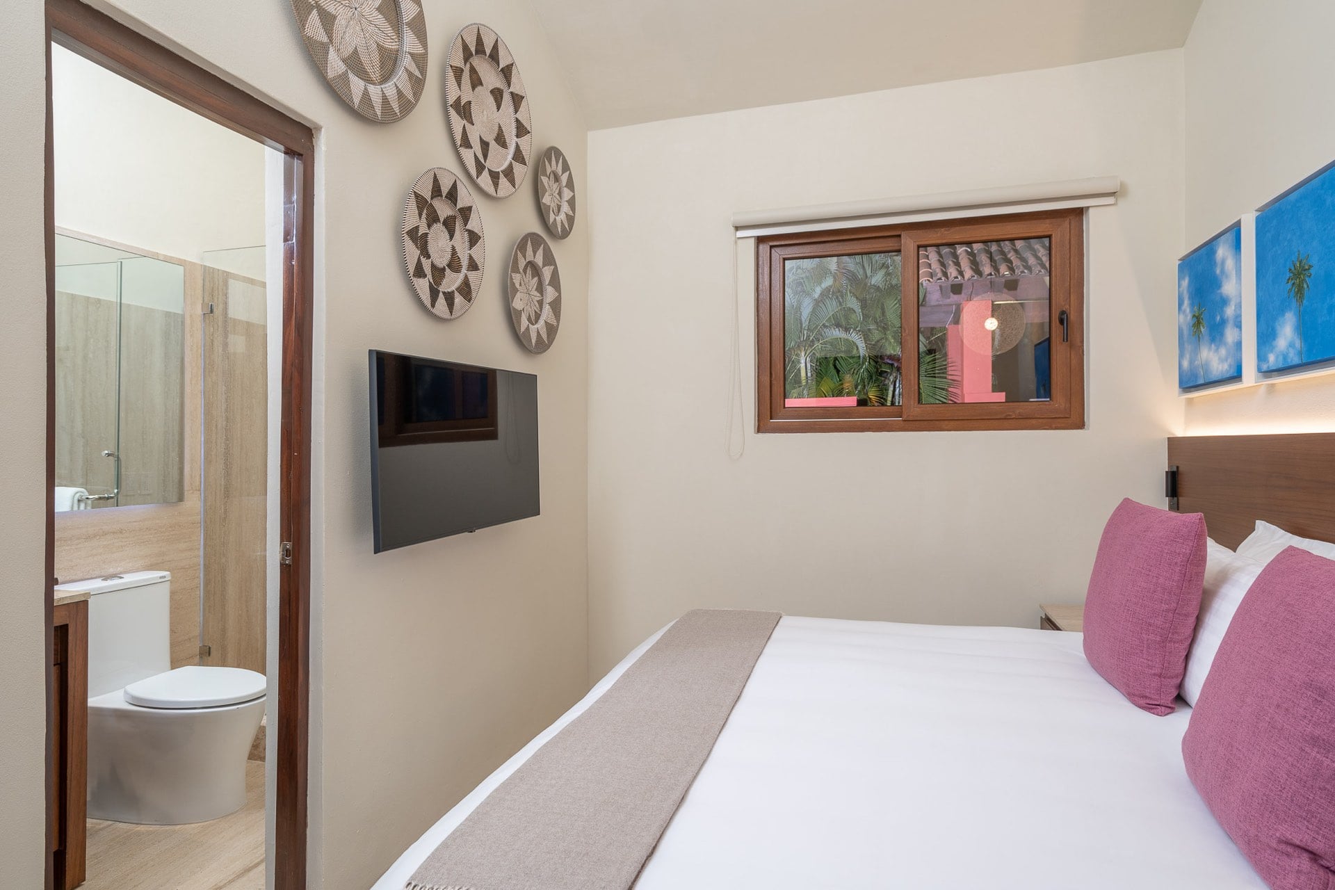 Guest suite king bed with rose pillows, cluster of woven basket art above, and open ensuite with glass shower, Villa Pacífica, Ranchos Estates, Punta Mita.