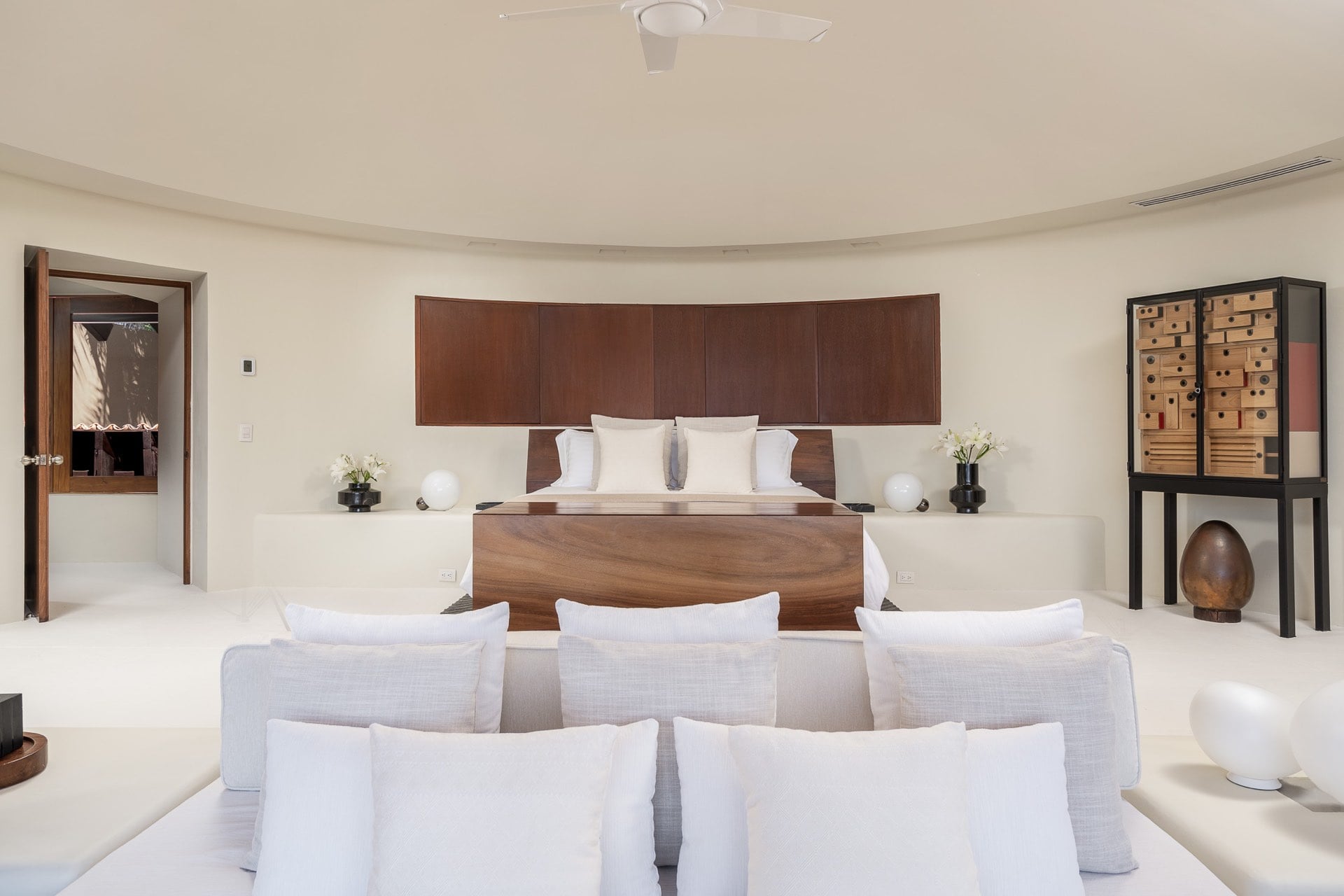 Primary suite from the foot of the bed showing curved walls, a sitting sofa, and an art cabinet beside the walnut headboard, Villa Pacífica, Ranchos Estates, Punta Mita.
