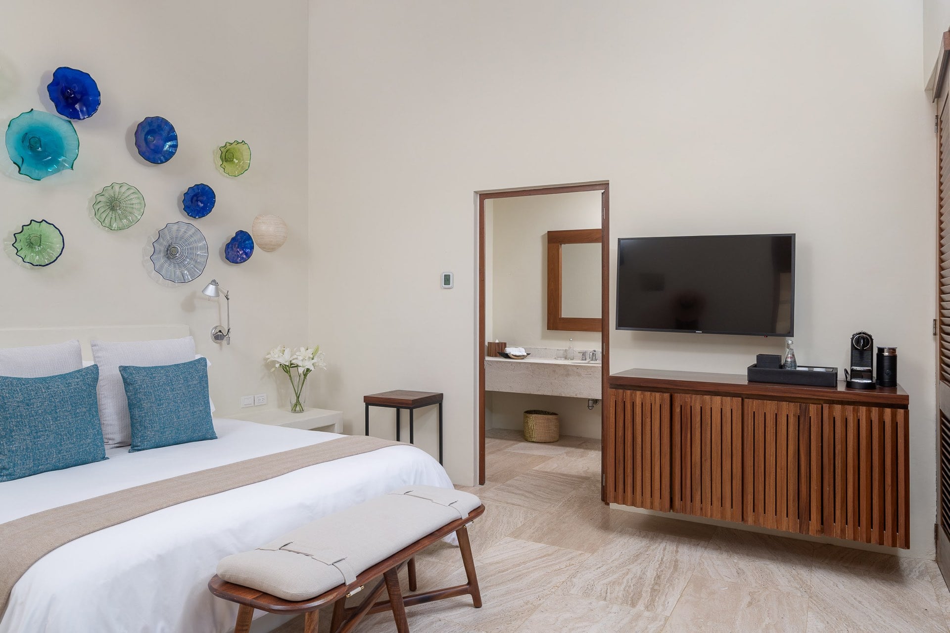 Guest suite king bed with blown-glass wall sculpture, wall-mounted TV, and open passage to travertine vanity, Villa Pacífica, Ranchos Estates, Punta Mita.