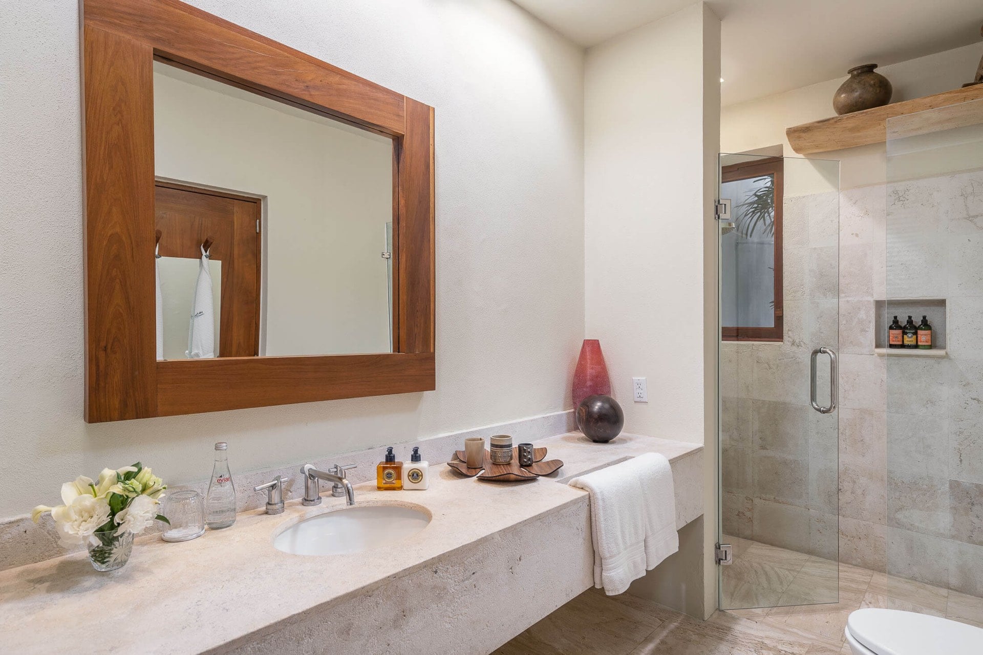 Ensuite bath with travertine vanity, teak-framed mirror, and glass-enclosed shower with niche toiletries, Villa Pacífica, Ranchos Estates, Punta Mita.