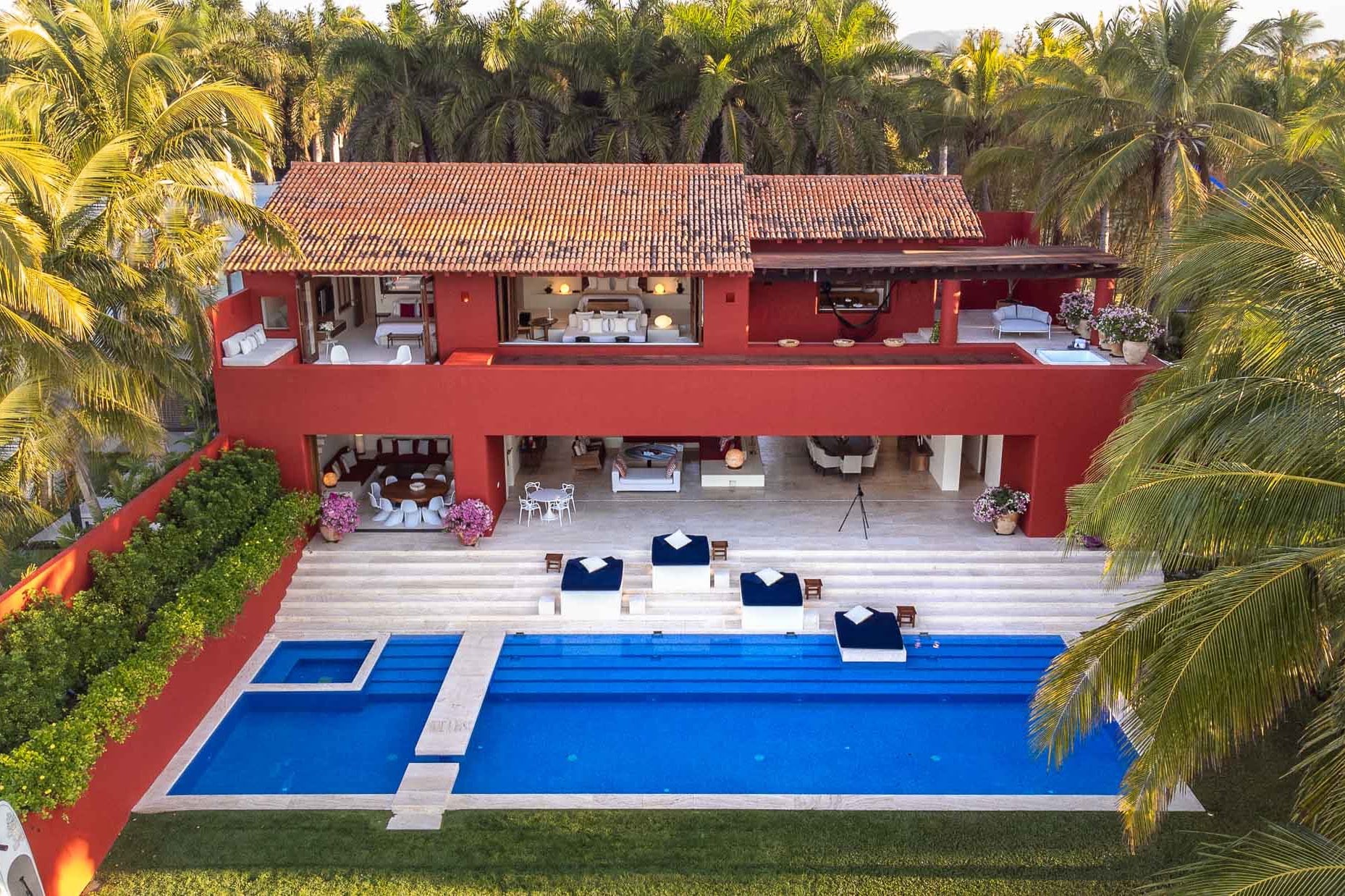 Aerial view of the red villa roof, lap pool with navy daybeds, and open living pavilion surrounded by palms, Villa Pacífica, Ranchos Estates, Punta Mita.