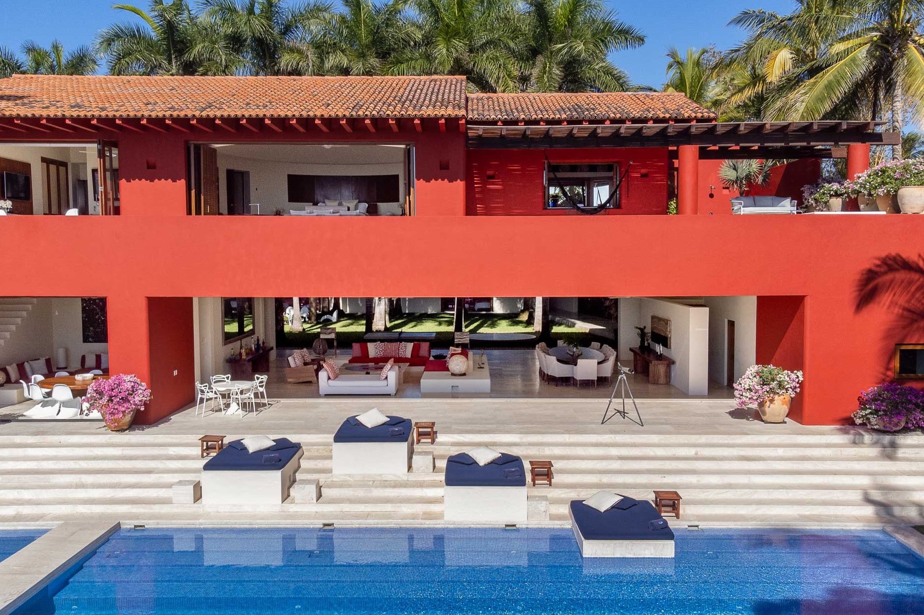 Villa facade from pool level showing open living pavilion with sofas, travertine terrace, and navy pool daybeds, Villa Pacífica, Ranchos Estates, Punta Mita.