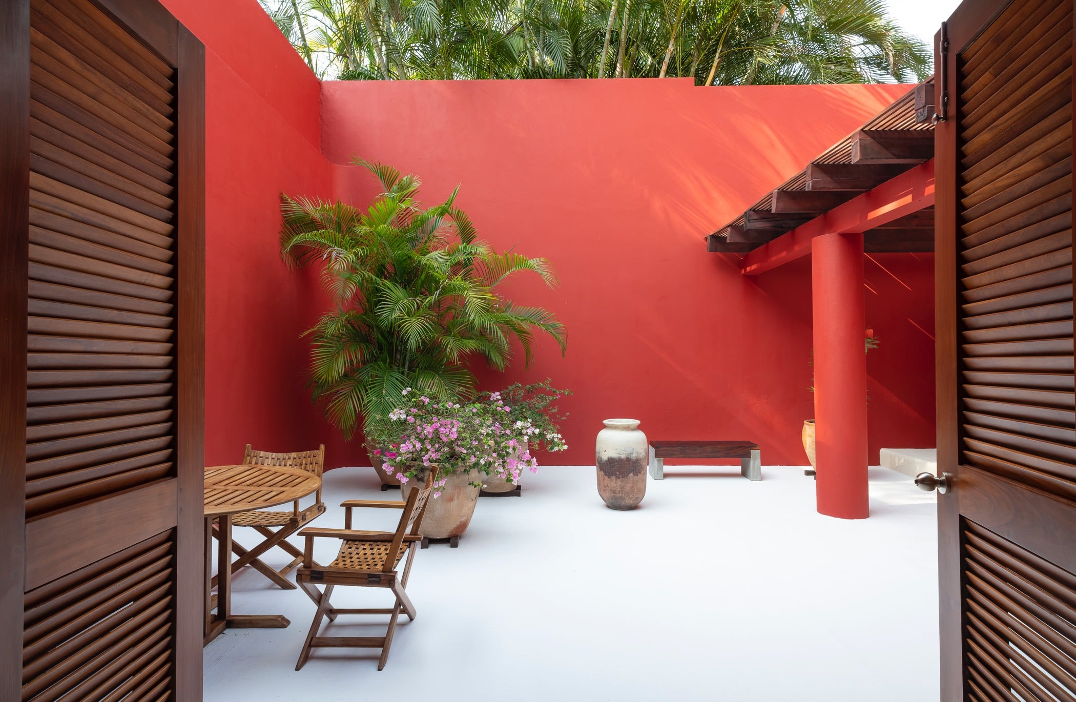 Private interior courtyard with red walls, teak bistro table, and a tall potted areca palm beneath the pergola, Villa Pacífica, Ranchos Estates, Punta Mita.