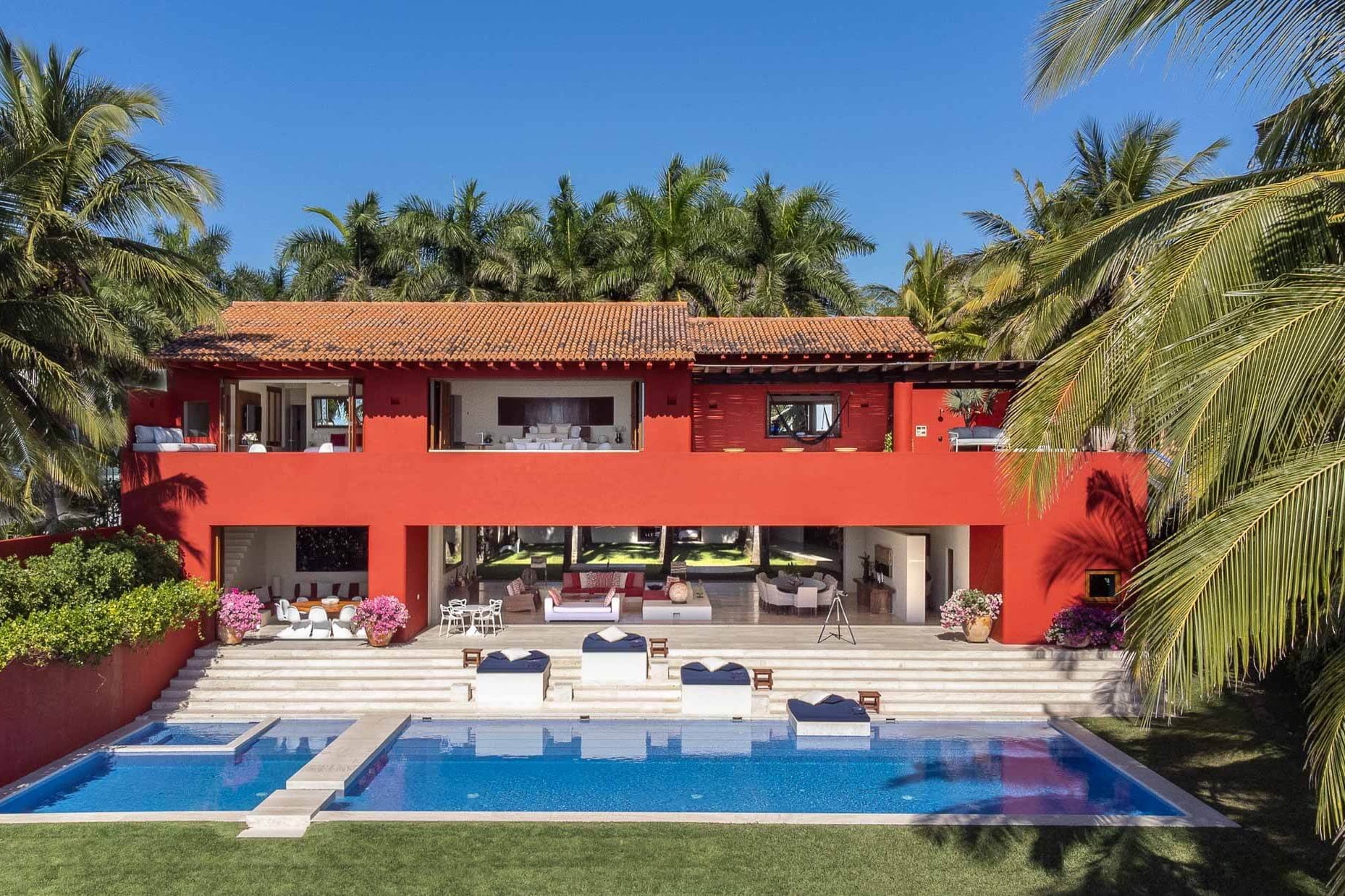 Daytime wide-angle of the red villa with full lap pool, travertine steps, and palm-framed lawn, Villa Pacífica, Ranchos Estates, Punta Mita.