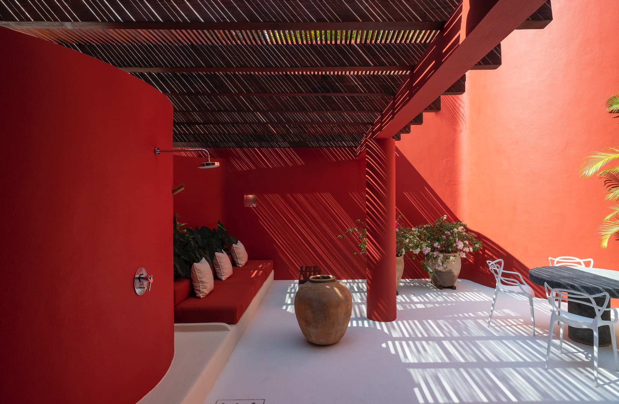 Outdoor spa courtyard with red pergola casting stripe shadows, wall shower head, built-in bench, and garden dining table, Villa Pacífica, Ranchos Estates, Punta Mita.