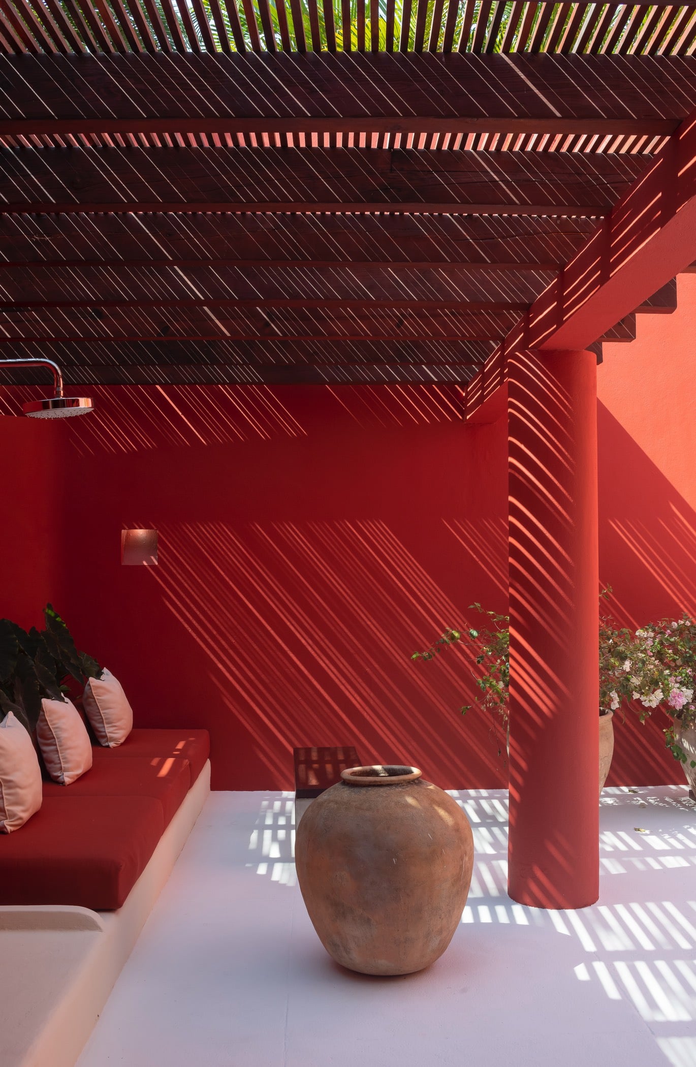 Outdoor rain shower head mounted on a crimson wall beneath a slatted wood pergola, with a terracotta urn and bougainvillea at the edge, Villa Pacífica, Ranchos Estates, Punta Mita.