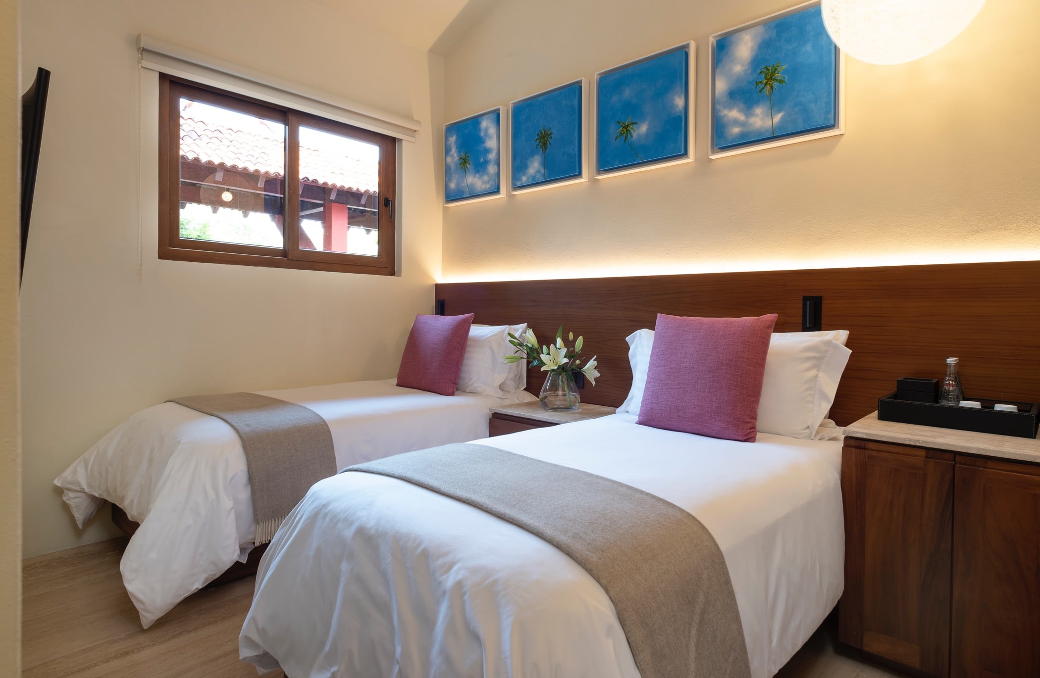 Twin-bed guest suite with a row of four palm-and-sky framed prints above the teak headboard, warm wall lighting, and a flat-screen TV, Villa Pacífica, Ranchos Estates, Punta Mita.