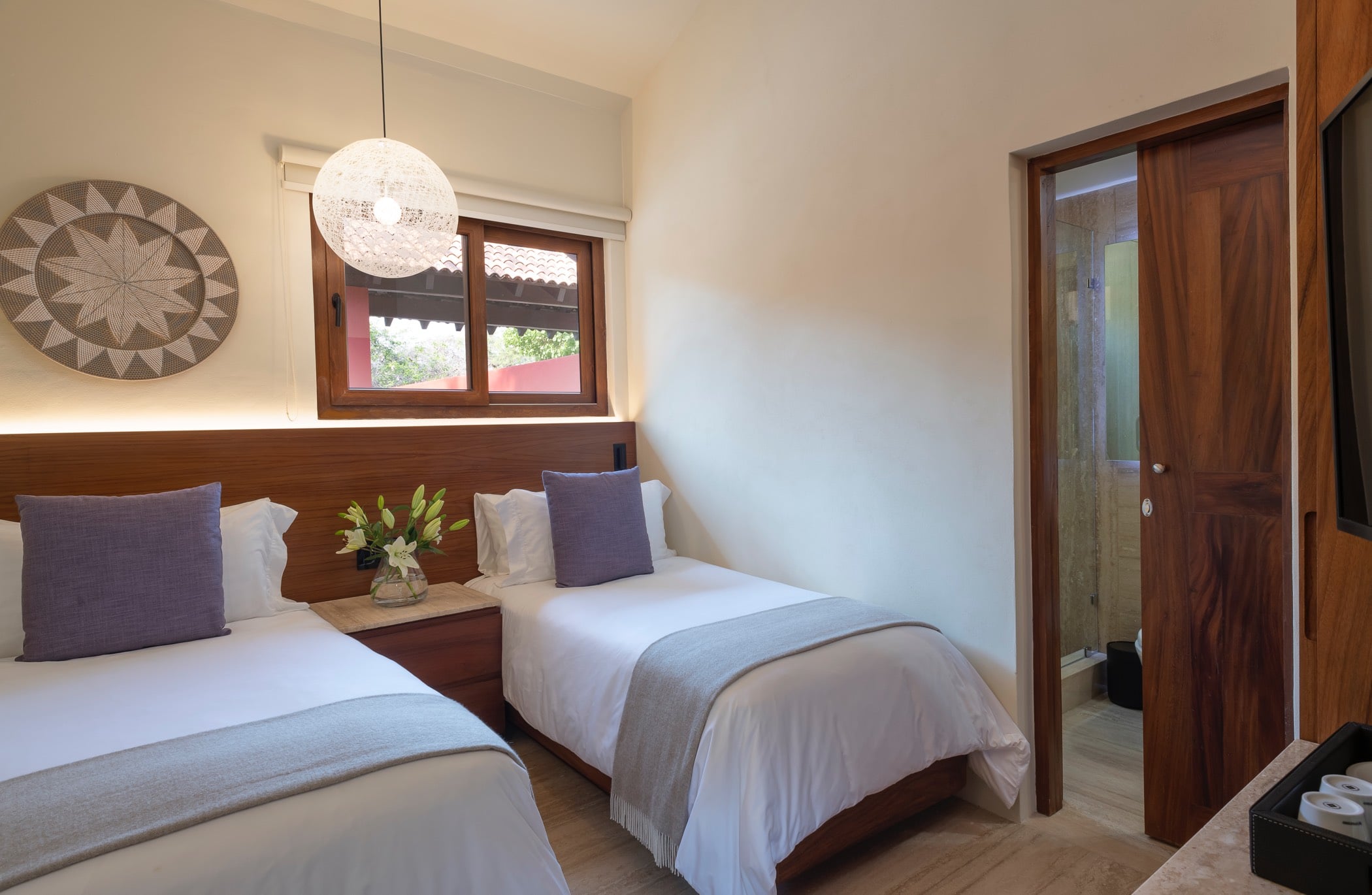 Twin-bed guest suite with woven basket wall art, a globe pendant light, lavender accent pillows, and a garden window, Villa Pacífica, Ranchos Estates, Punta Mita.