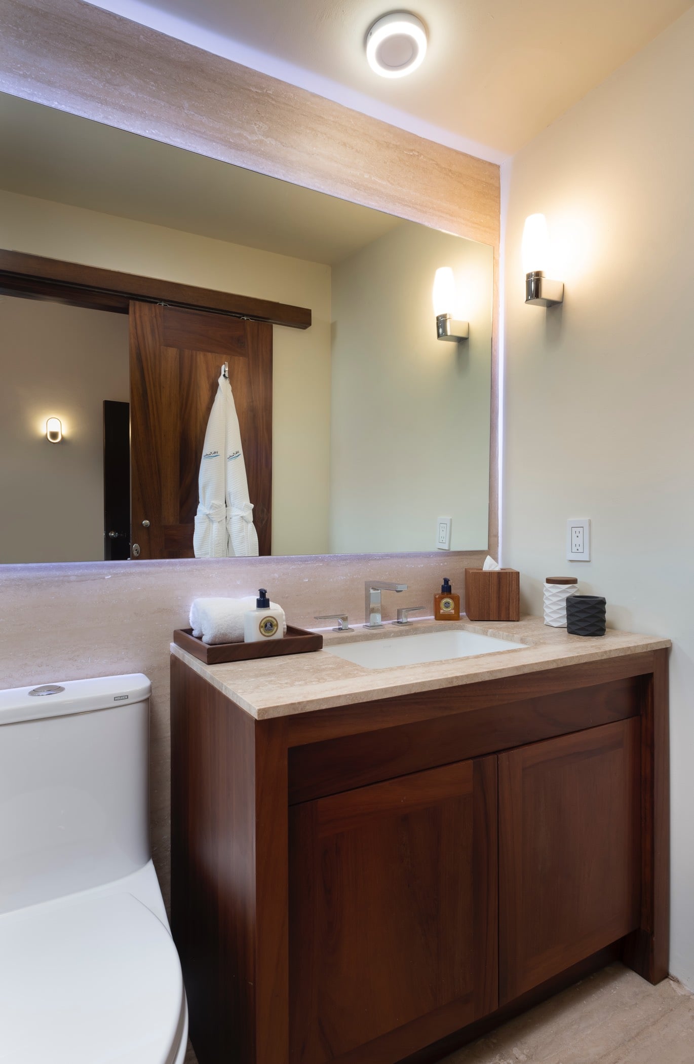 Single-vanity ensuite bathroom with teak cabinet, travertine countertop, large mirror, and robes reflected in the glass, Villa Pacífica, Ranchos Estates, Punta Mita.