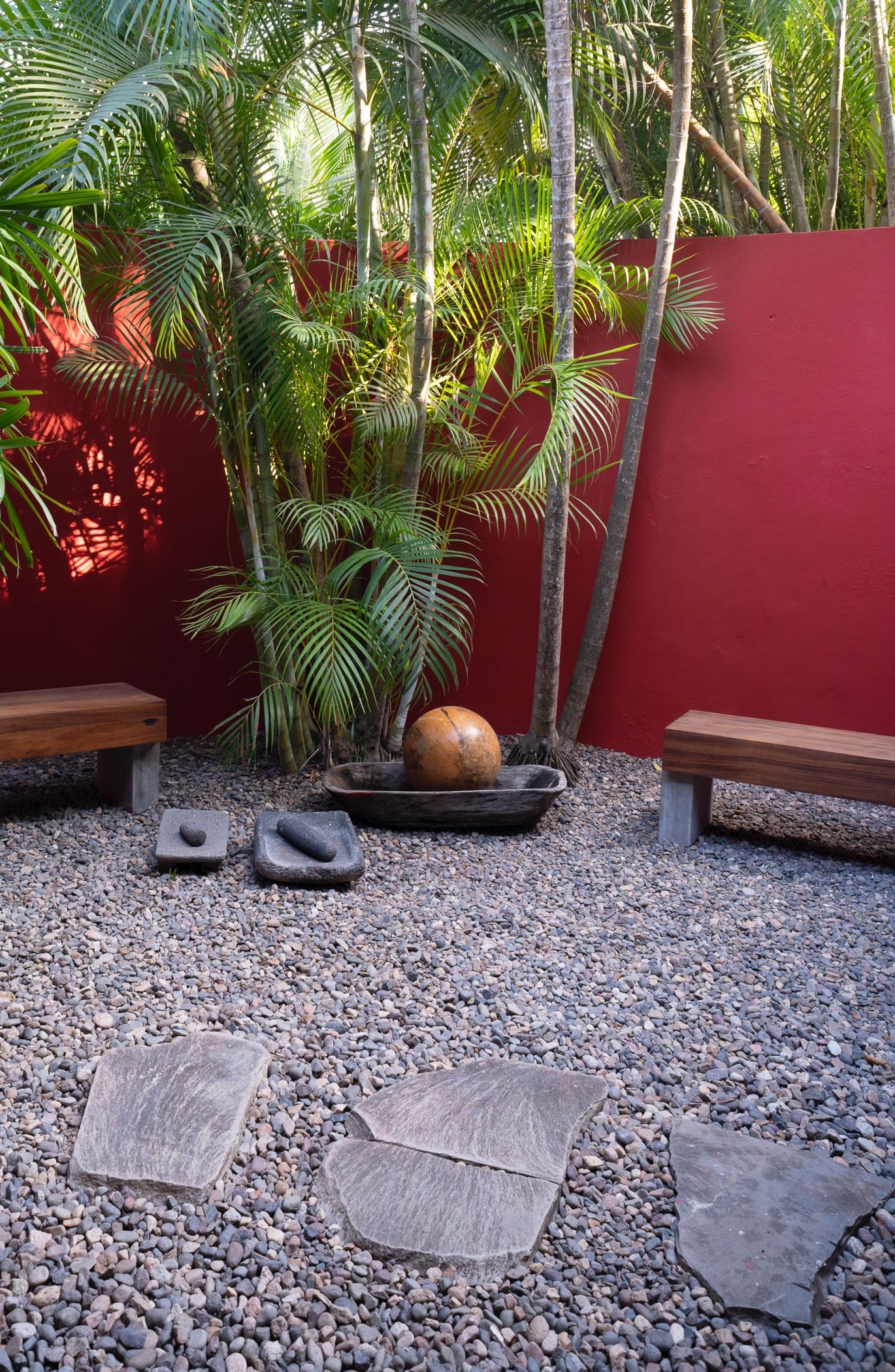 Gravel meditation garden with flat stone stepping pads, a carved wooden bowl, two teak benches, and palms rising behind a red boundary wall, Villa Pacífica, Ranchos Estates, Punta Mita.