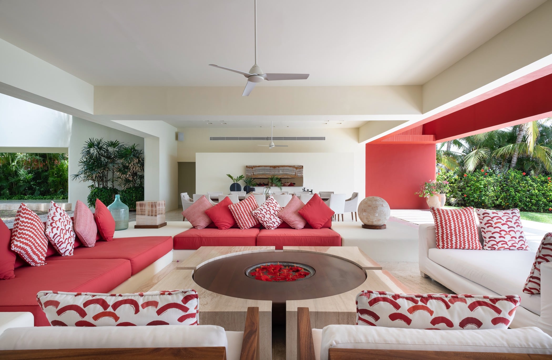 Open-air living pavilion with deep red sectional sofas, a round fire table, ceiling fans, dining table beyond, and palms framing the garden edge, Villa Pacífica, Ranchos Estates, Punta Mita.