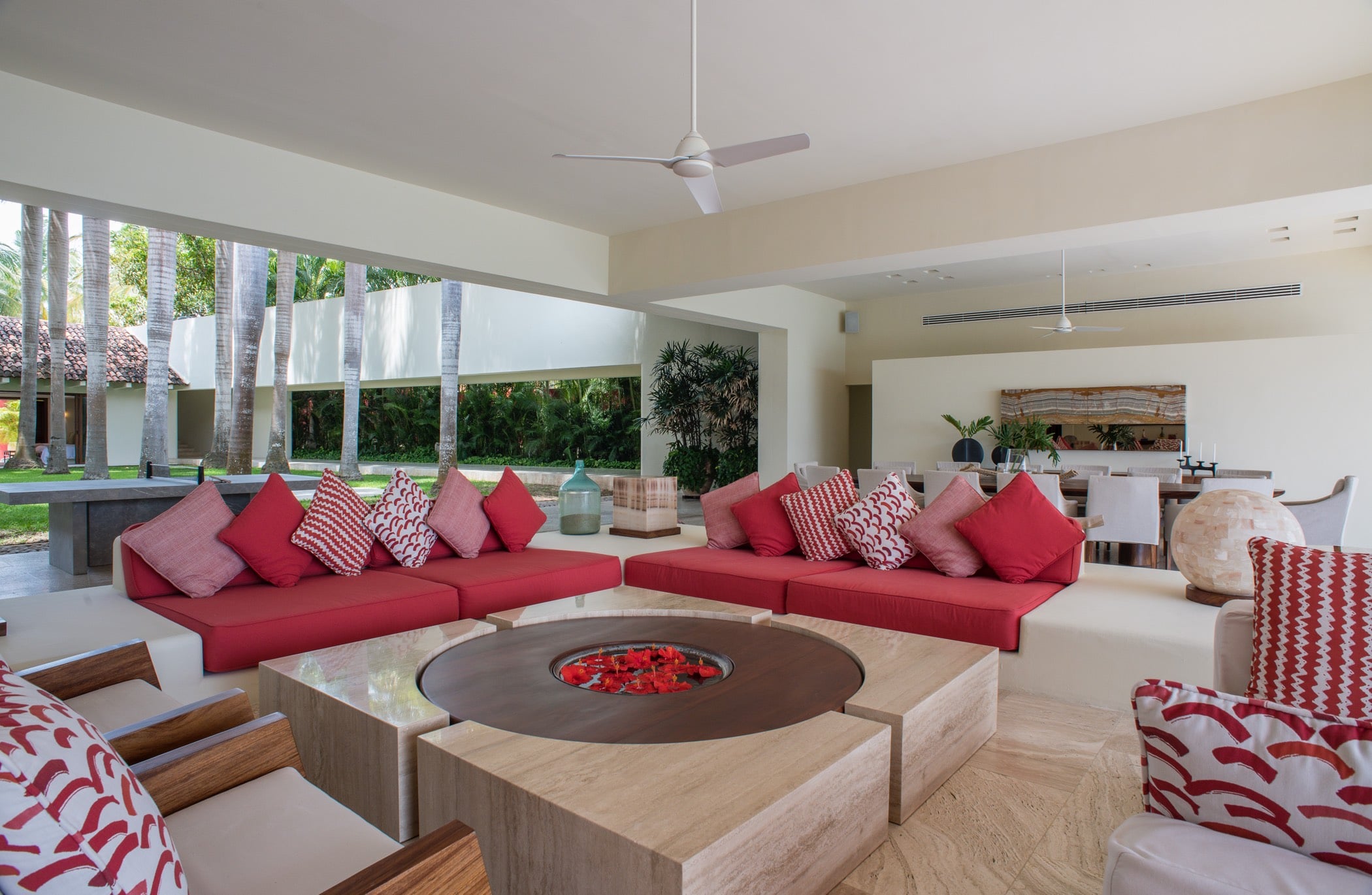 Open-air living pavilion showing red low-slung sofas around a travertine fire table, palm trunks visible through the open perimeter, dining chairs in the background, Villa Pacífica, Ranchos Estates, Punta Mita.