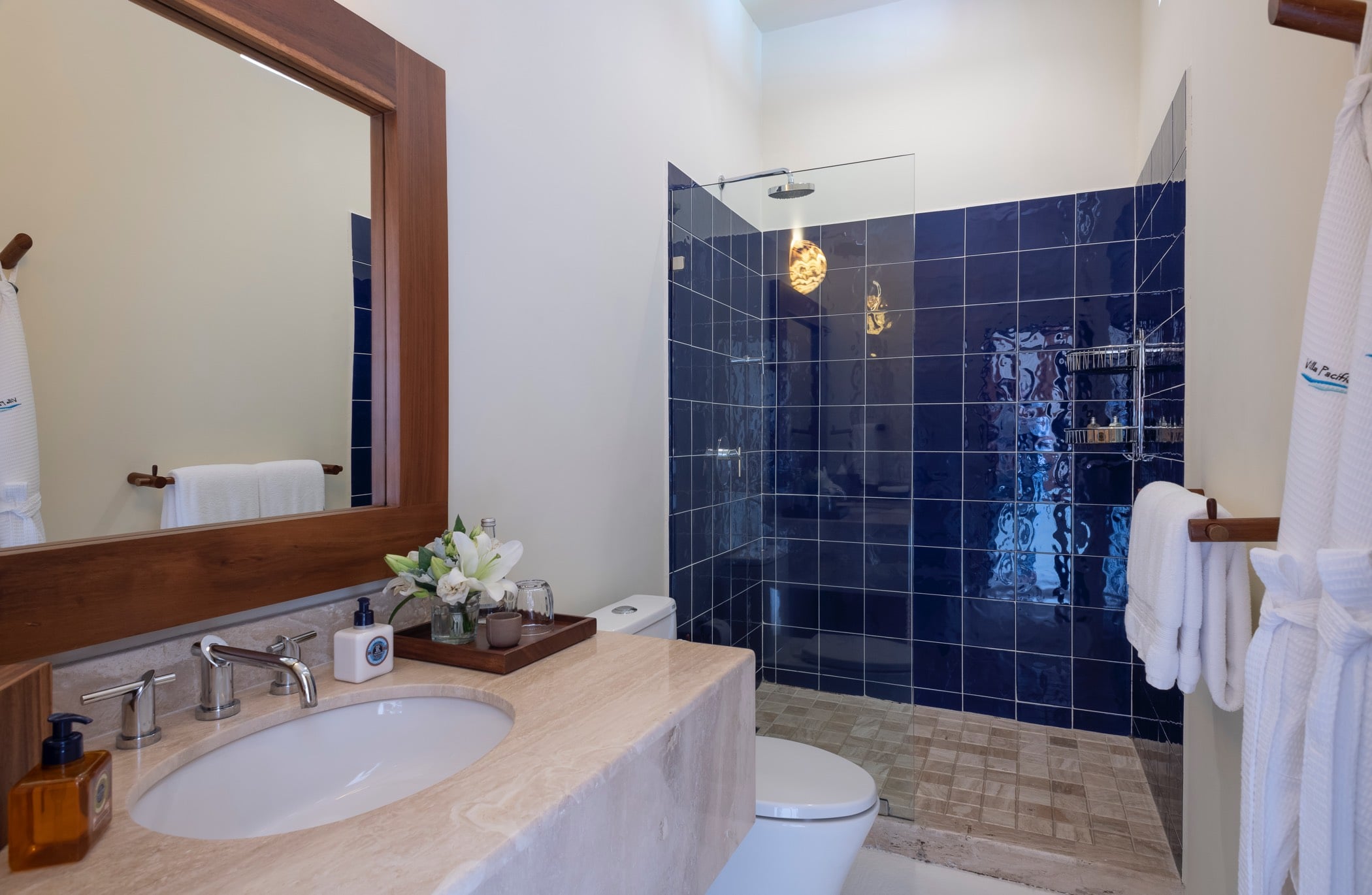 Ensuite bathroom with glossy cobalt-blue tiled shower, teak-framed mirror, marble vanity, and Villa Pacífica branded robes on a wooden rack, Villa Pacífica, Ranchos Estates, Punta Mita.