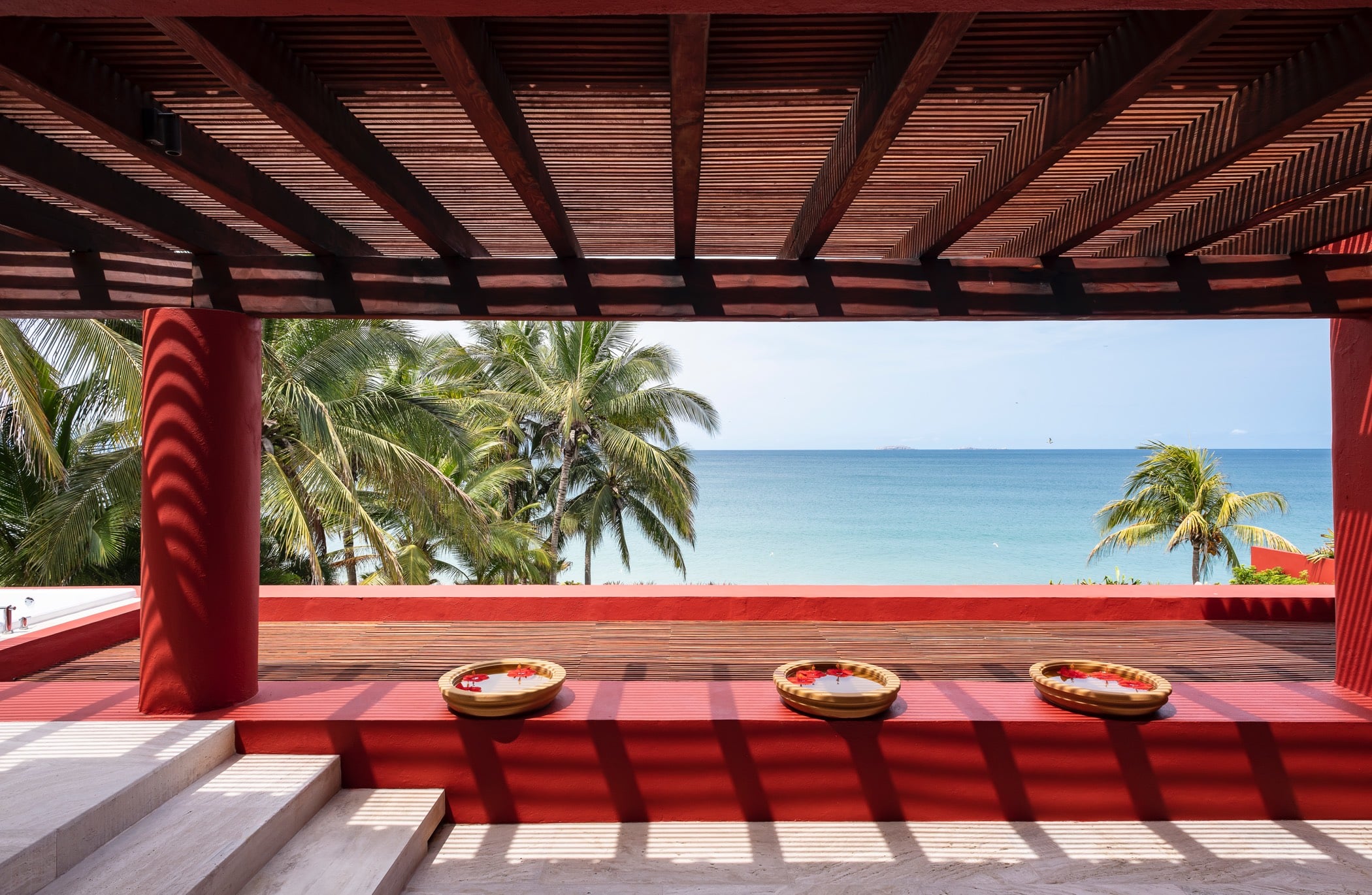 View from beneath a red pergola looking over a low terrace ledge to palms and the turquoise Pacific Ocean, with three woven bowls on the ledge, Villa Pacífica, Ranchos Estates, Punta Mita.