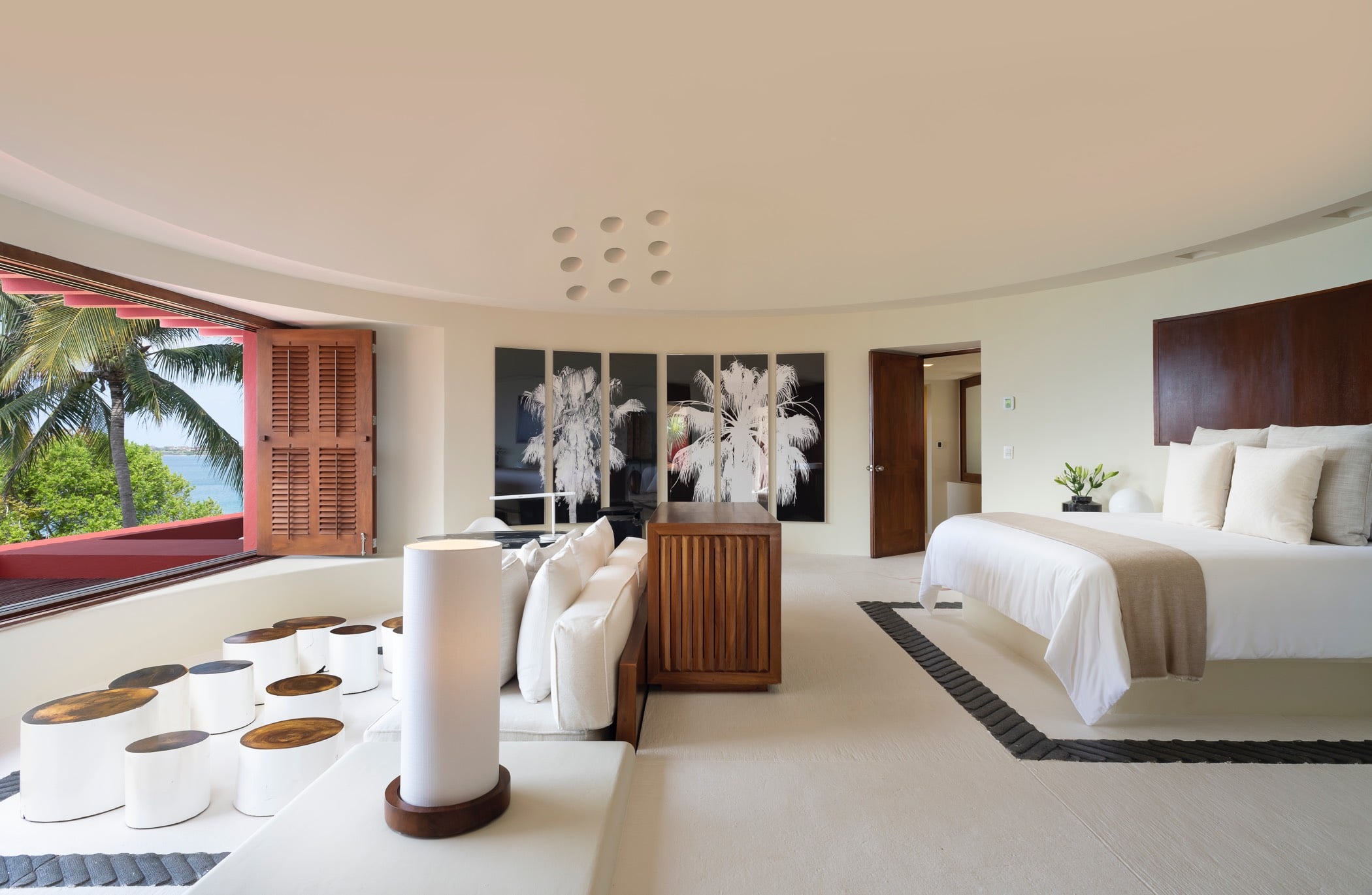 Primary suite with a curved white ceiling, king bed, teak-console sofa, large black-and-white palm art panels, and shuttered wooden doors opening to an ocean view, Villa Pacífica, Ranchos Estates, Punta Mita.