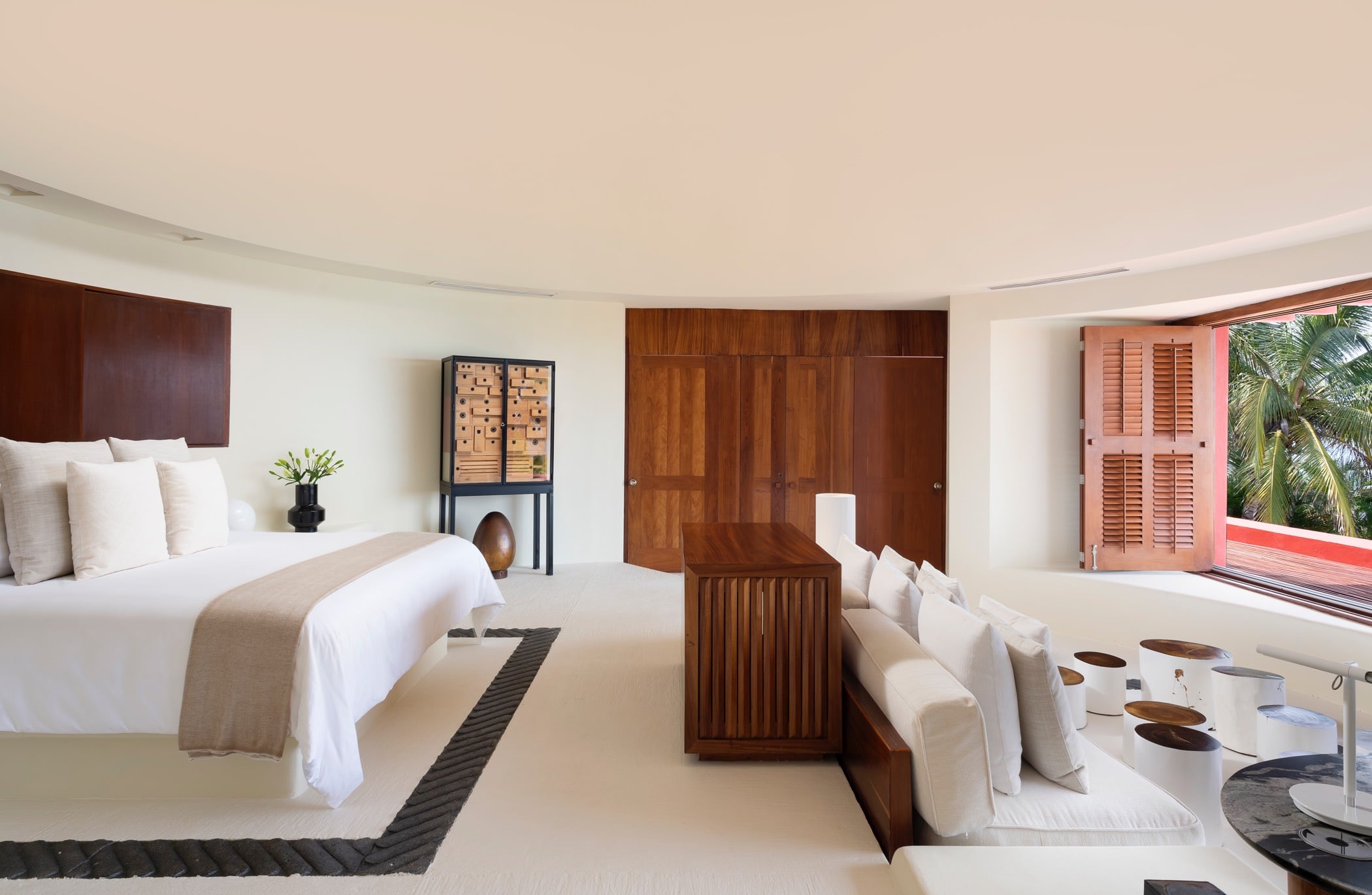 Primary suite with king bed, full-height teak wardrobe wall, a collector-cabinet piece, and shuttered windows overlooking garden palms, Villa Pacífica, Ranchos Estates, Punta Mita.