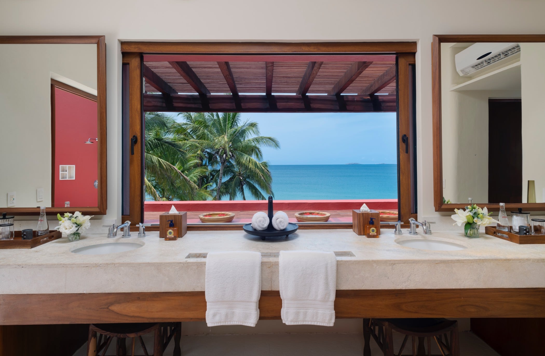 Primary suite double vanity centered on an open window framing palm fronds and the turquoise ocean, teak mirrors flanking each side, Villa Pacífica, Ranchos Estates, Punta Mita.