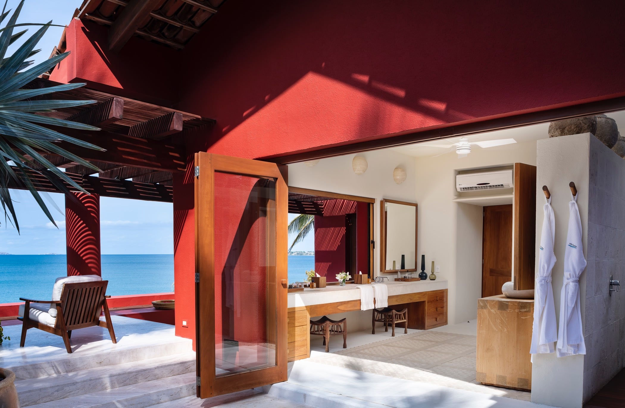 Primary suite bathroom seen through an open pivot door, double vanity with teak mirrors and robe hooks visible, the red terrace and Pacific Ocean framed in the background, Villa Pacífica, Ranchos Estates, Punta Mita.