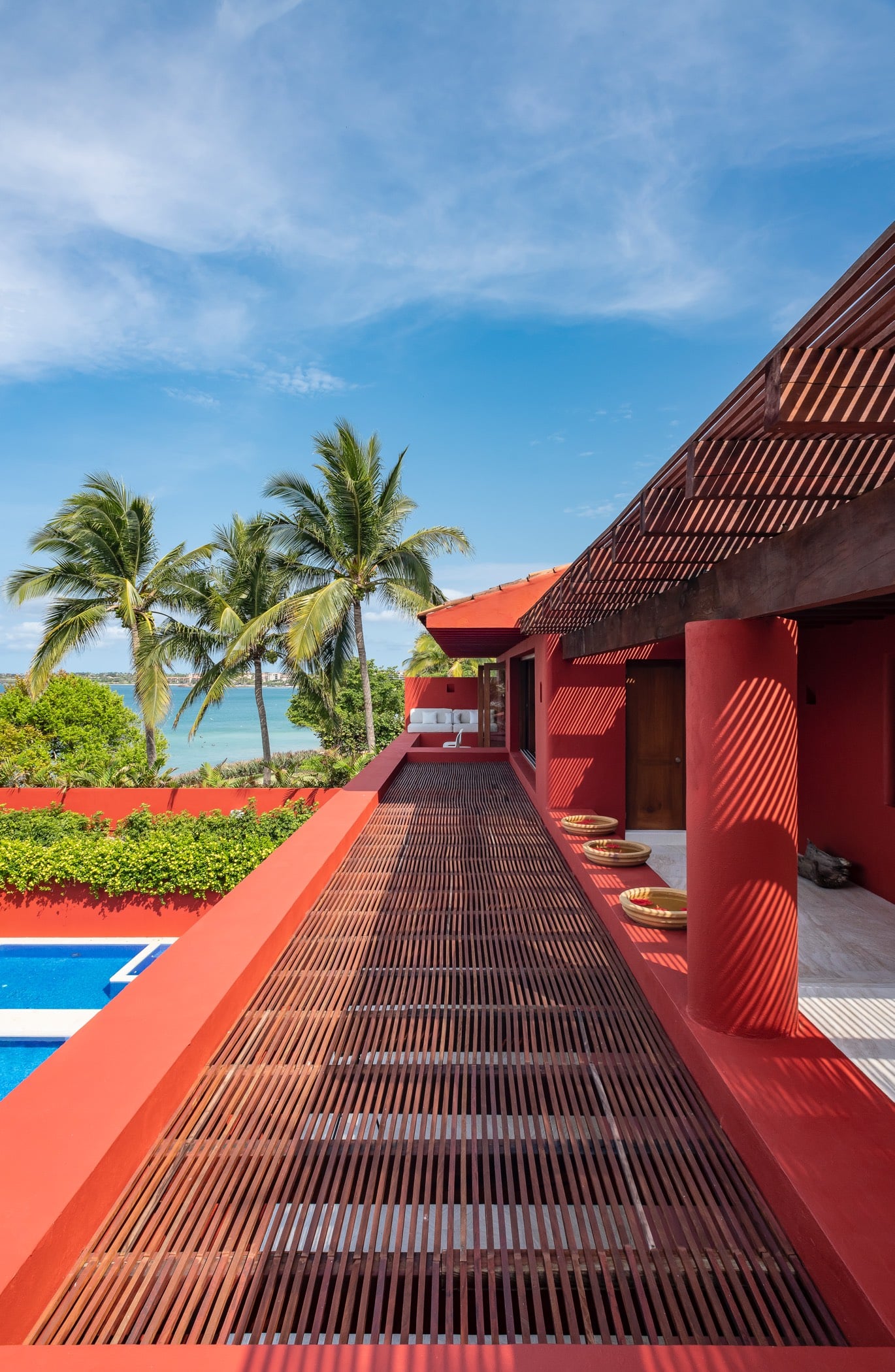 Elevated pool deck walkway with slatted wood decking, pool edge at left, red parapets, tall palms, and the ocean visible under a blue sky, Villa Pacífica, Ranchos Estates, Punta Mita.