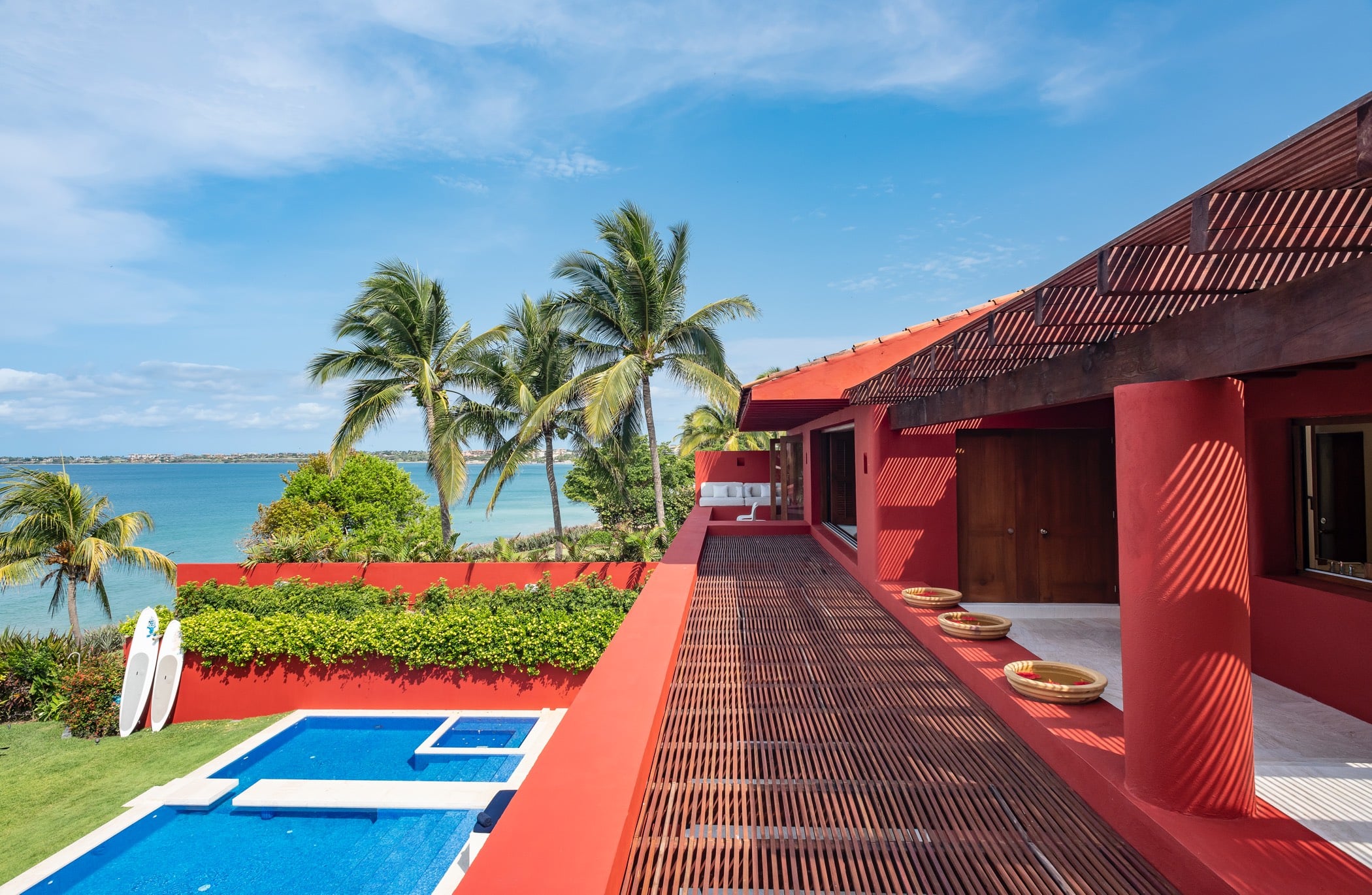 Exterior view of pool, lawn with surfboards, red facade, slatted wood pergola walkway, and the Pacific Ocean under open sky, Villa Pacífica, Ranchos Estates, Punta Mita.