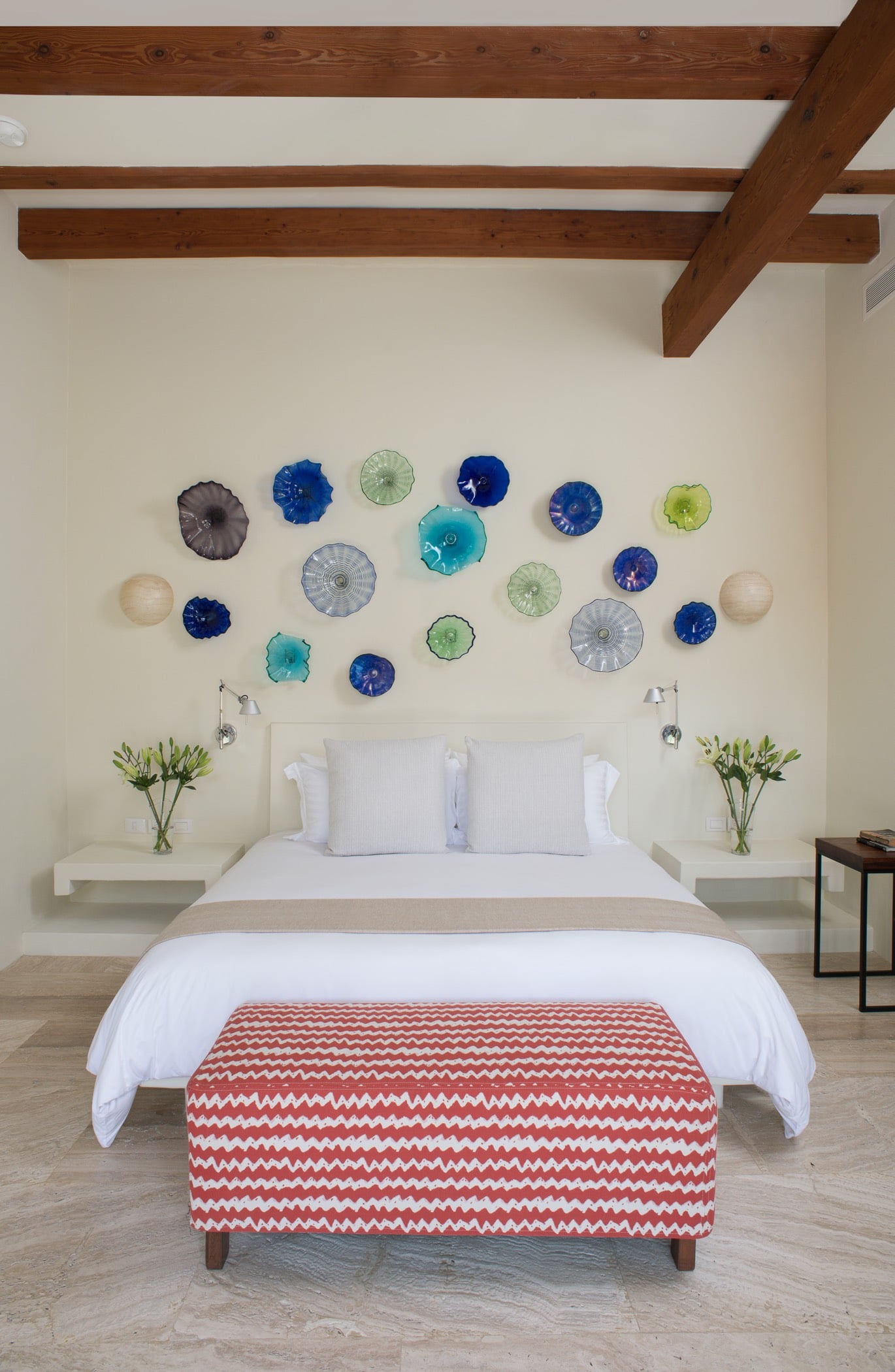 Guest suite with king bed, a cluster of colorful blown-glass disc art above the headboard, timber ceiling beams, and a patterned bench at the foot, Villa Pacífica, Ranchos Estates, Punta Mita.