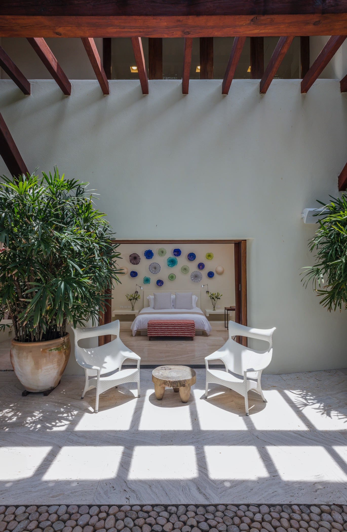 Private terrace outside guest suite with two wicker armchairs and a stump table, large palms in terracotta pots, timber-beam ceiling, and the glass-art suite visible through an open door, Villa Pacífica, Ranchos Estates, Punta Mita.