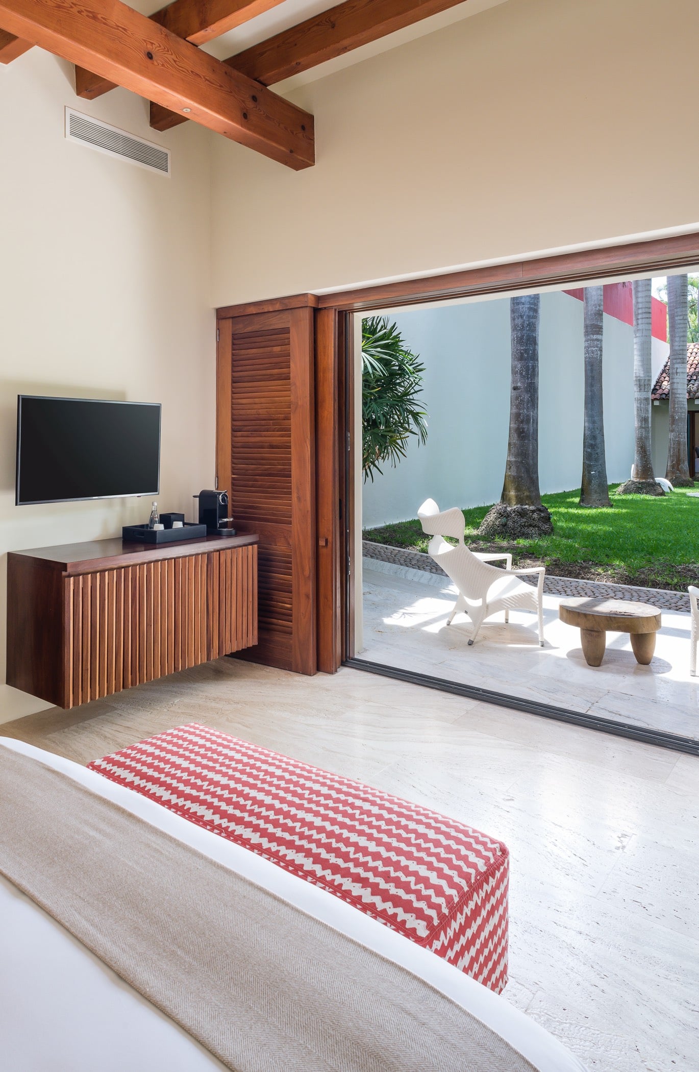 Guest suite with king bed, slatted teak TV cabinet, flat-screen TV, and sliding glass doors fully open to a garden terrace with palm trunks, Villa Pacífica, Ranchos Estates, Punta Mita.