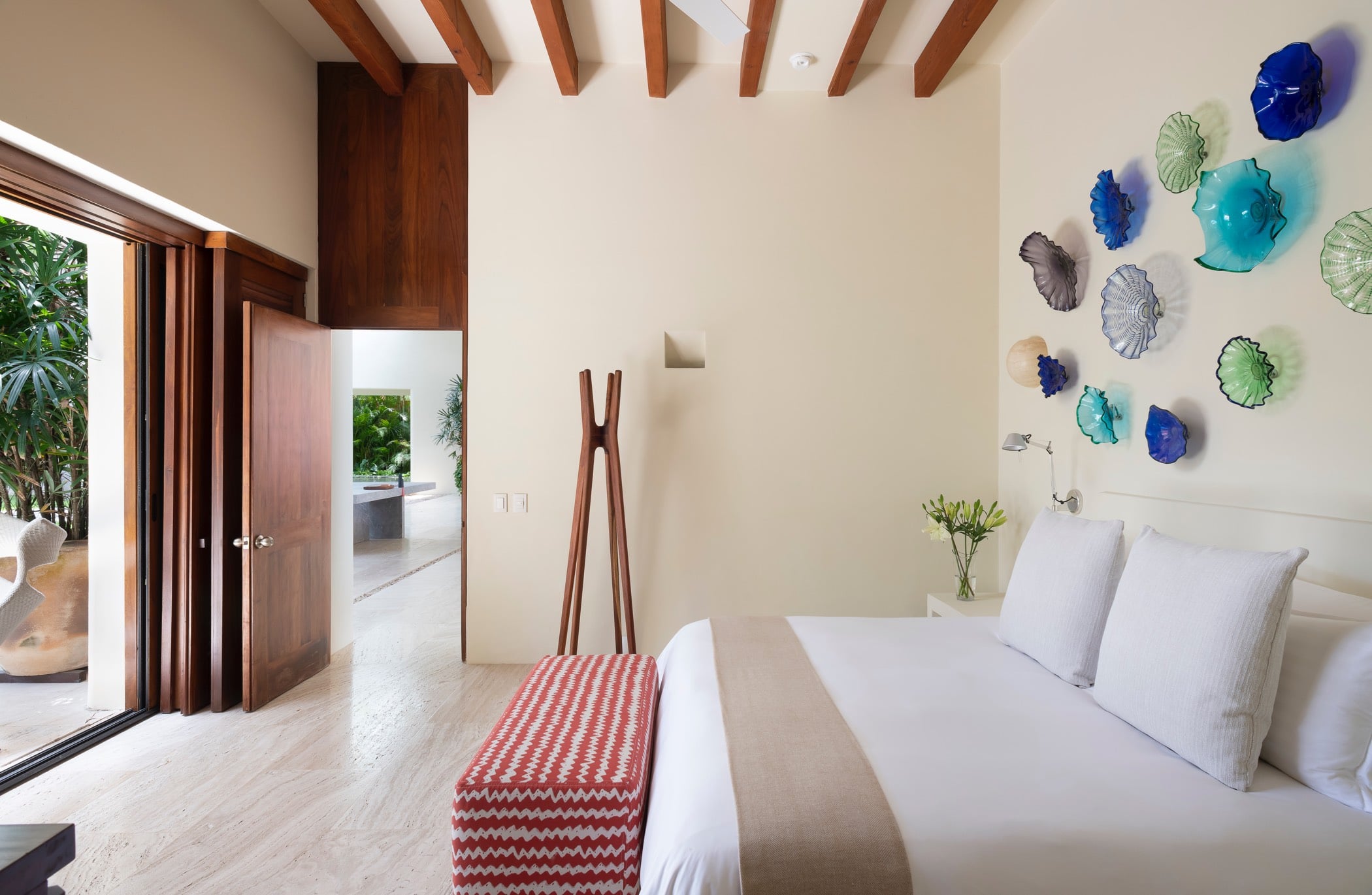 Guest suite with folding wood doors open, blown-glass disc wall art visible at right, a sculptural teak coat stand, and a glimpse of the ensuite bathroom beyond, Villa Pacífica, Ranchos Estates, Punta Mita.
