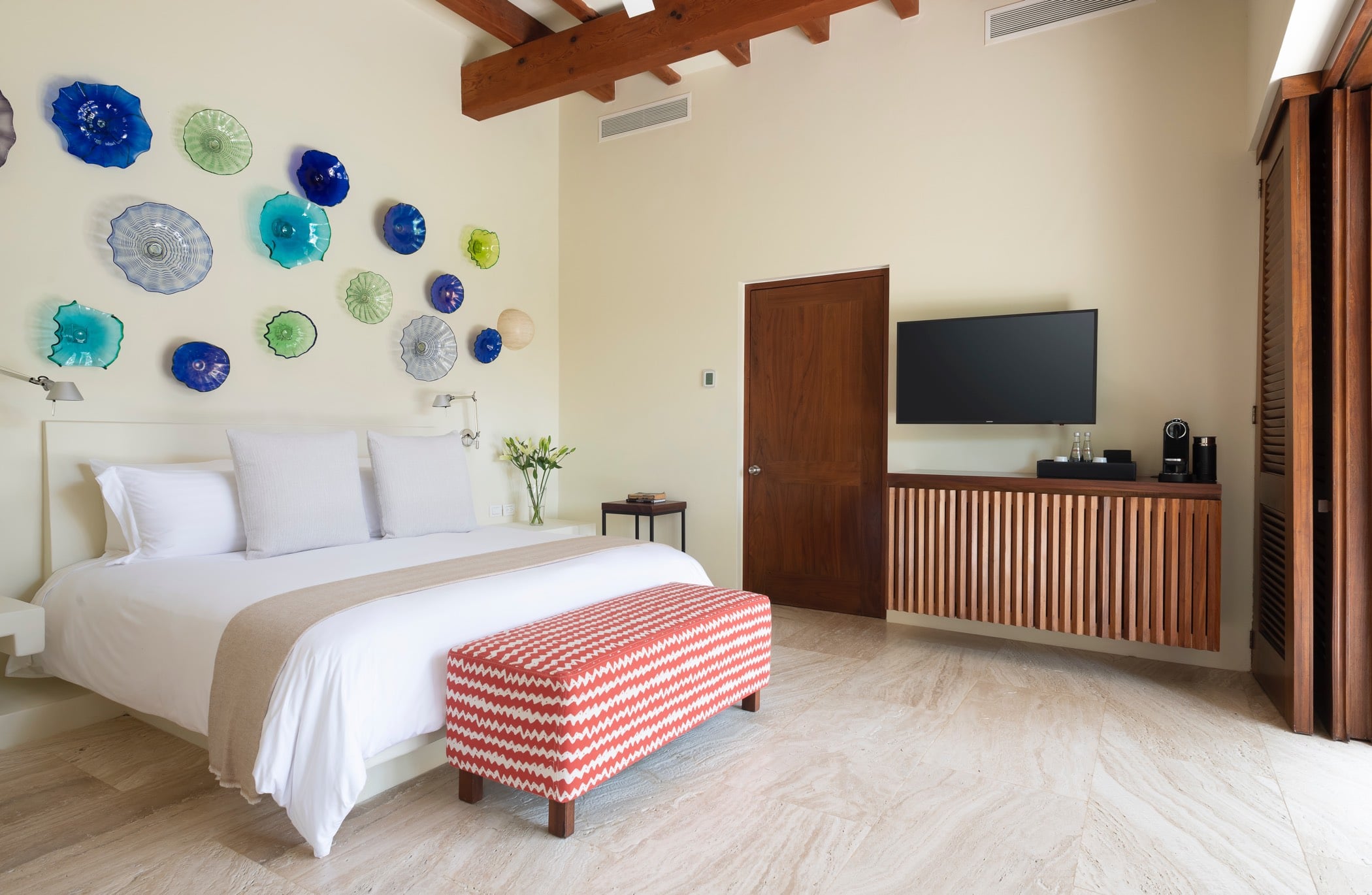 Guest suite full-room view with king bed, blown-glass disc art on the wall, teak slatted media console, and louvered shutters opening to the terrace, Villa Pacífica, Ranchos Estates, Punta Mita.