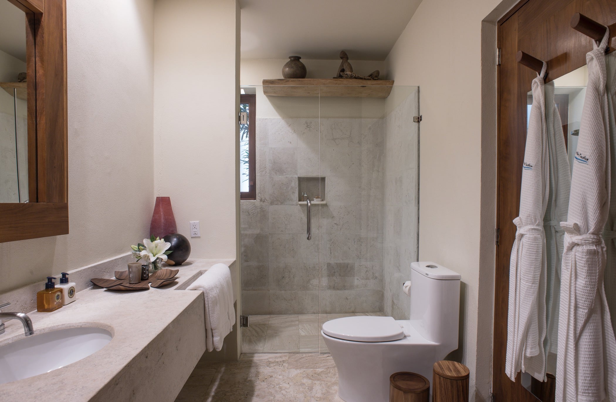 Guest ensuite bathroom with travertine-tiled glass shower, marble countertop, teak-framed mirror, and Villa Pacífica branded robes hanging by the door, Villa Pacífica, Ranchos Estates, Punta Mita.