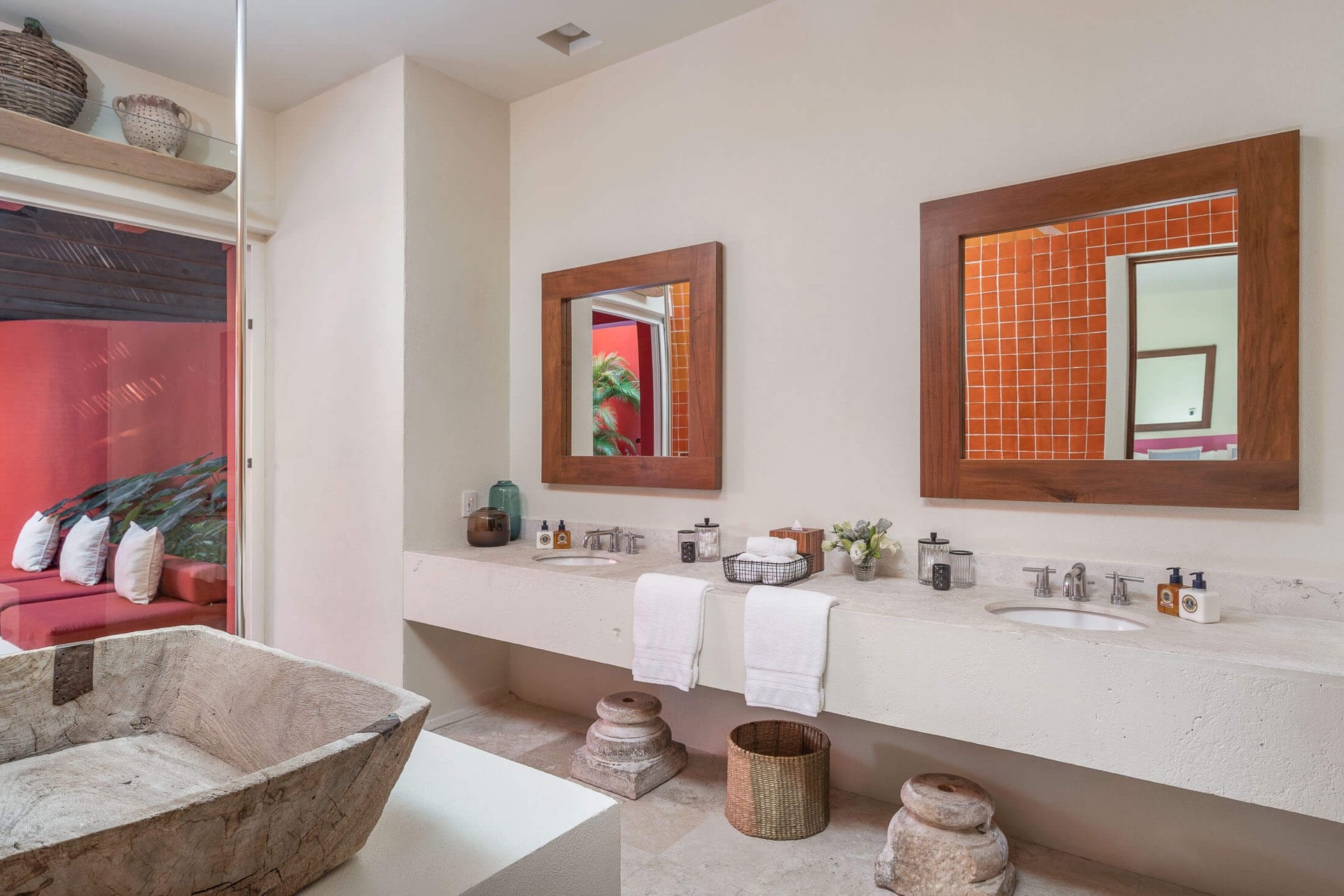 Double vanity with stone countertop and two wood-framed mirrors reflecting terracotta tile, carved stone soaking tub at left, open doorway to bedroom beyond, Villa Pacífica, Ranchos Estates, Punta Mita.