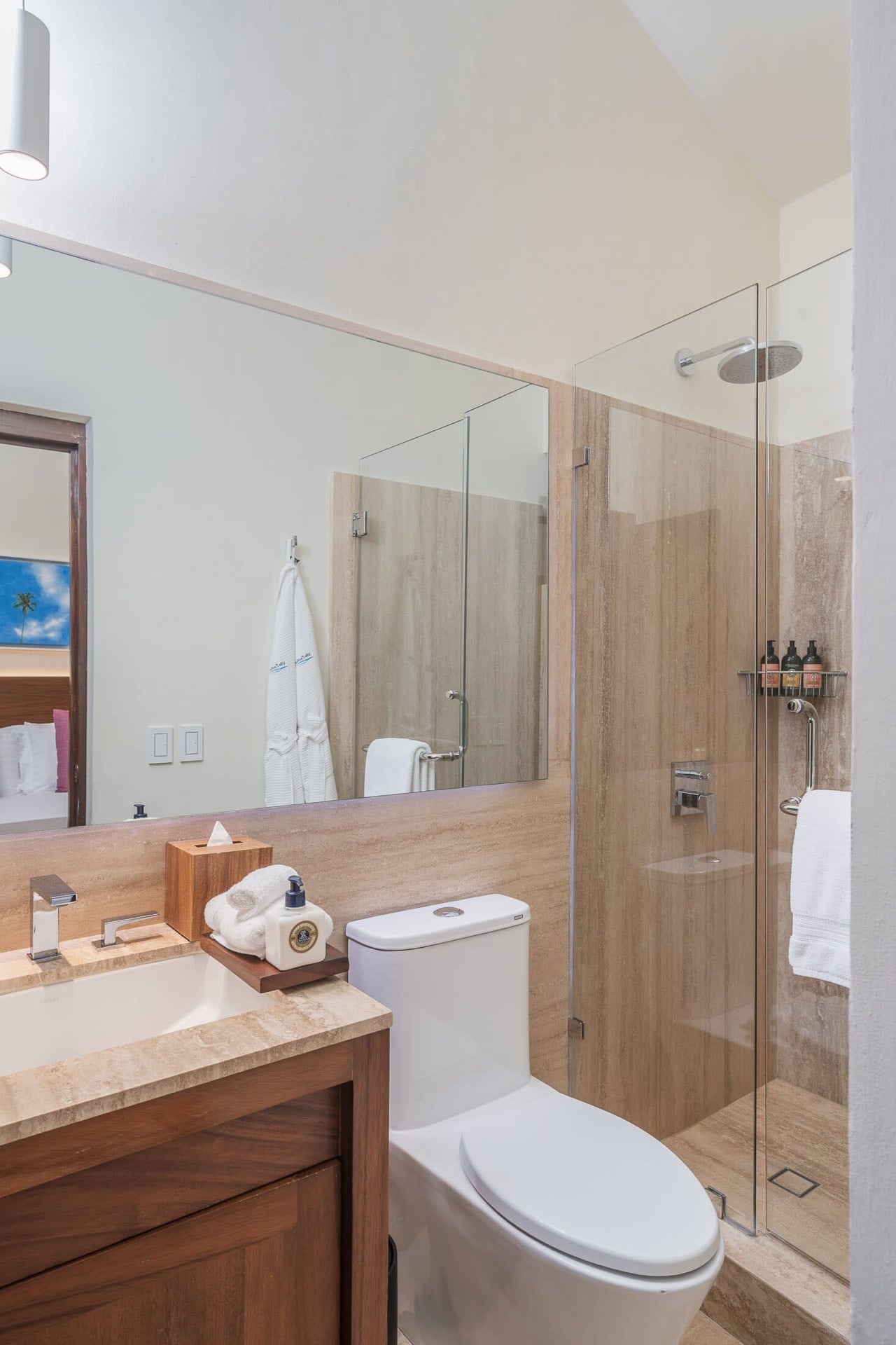 Travertine walk-in shower with glass partition and rain head beside a walnut vanity cabinet and stone countertop, bedroom visible through open door, Villa Pacífica, Ranchos Estates, Punta Mita.