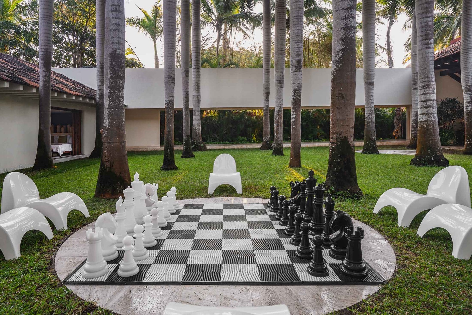 Oversized chess set on a circular stone platform amid a tall palm grove, white sculptural chairs around the board, villa pavilions in the background, Villa Pacífica, Ranchos Estates, Punta Mita.