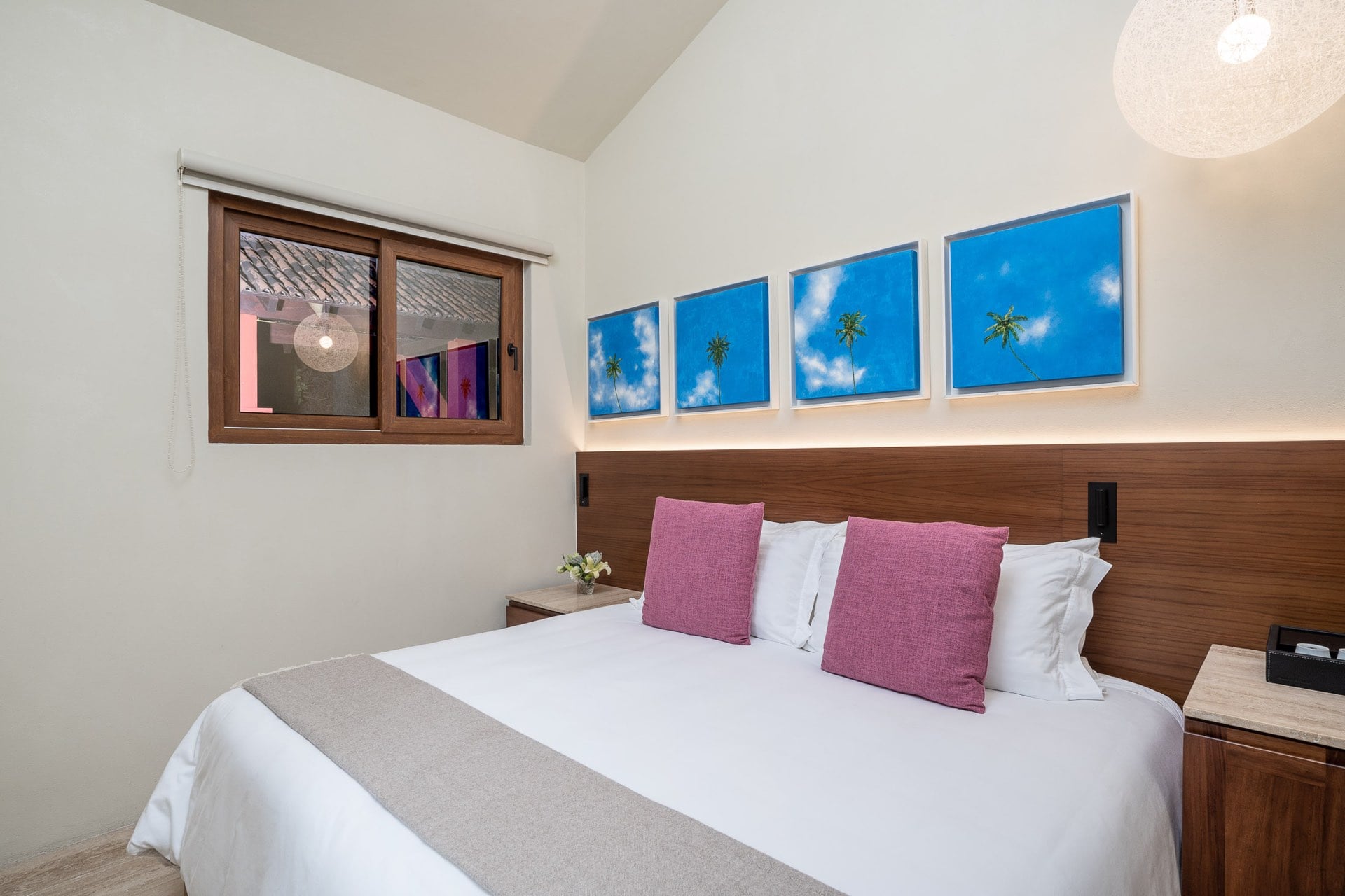 Guest suite queen bed with walnut headboard wall panel, four blue-sky palm photography panels above, rose-pink accent pillows, and globe pendant light, Villa Pacífica, Ranchos Estates, Punta Mita.