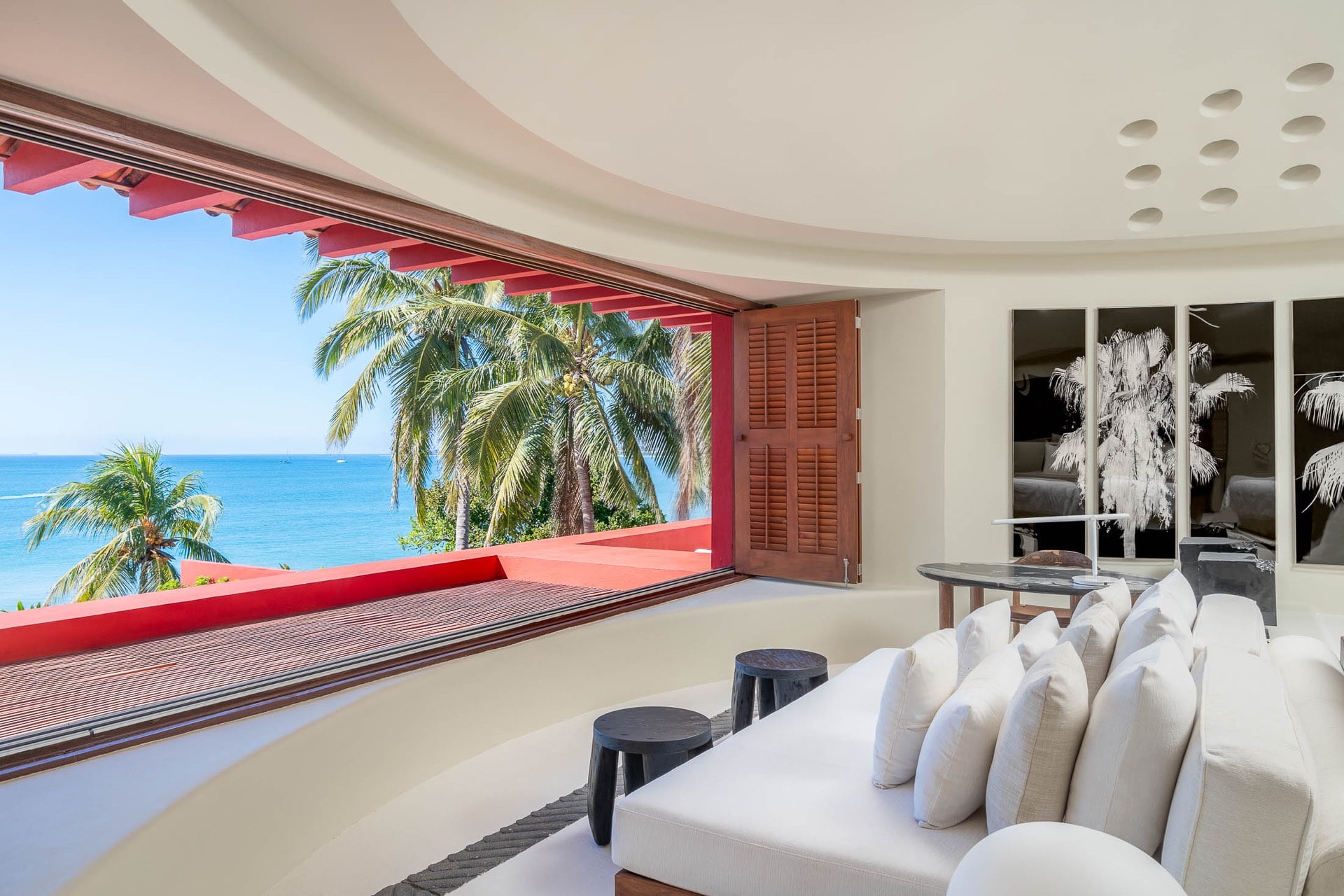 Primary suite sitting area with curved plaster ceiling and wide picture opening to the Pacific Ocean, coconut palms, and red terrace railing, white sofa and dark stools in foreground, Villa Pacífica, Ranchos Estates, Punta Mita.