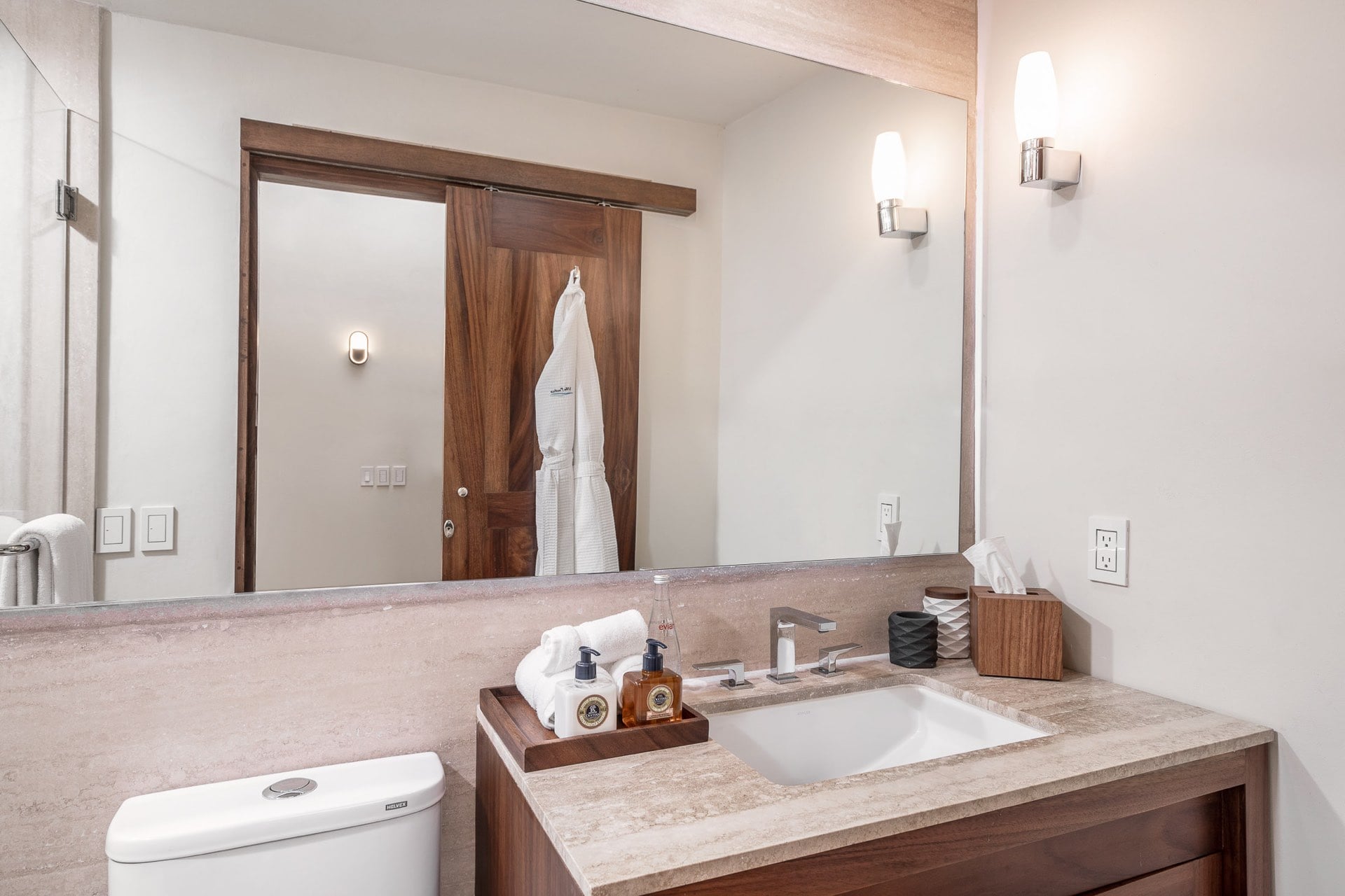 Single travertine-top vanity with walnut cabinet and large mirror reflecting a sliding barn-style wood door with hanging robe, wall sconce and L'Occitane amenities on tray, Villa Pacífica, Ranchos Estates, Punta Mita.