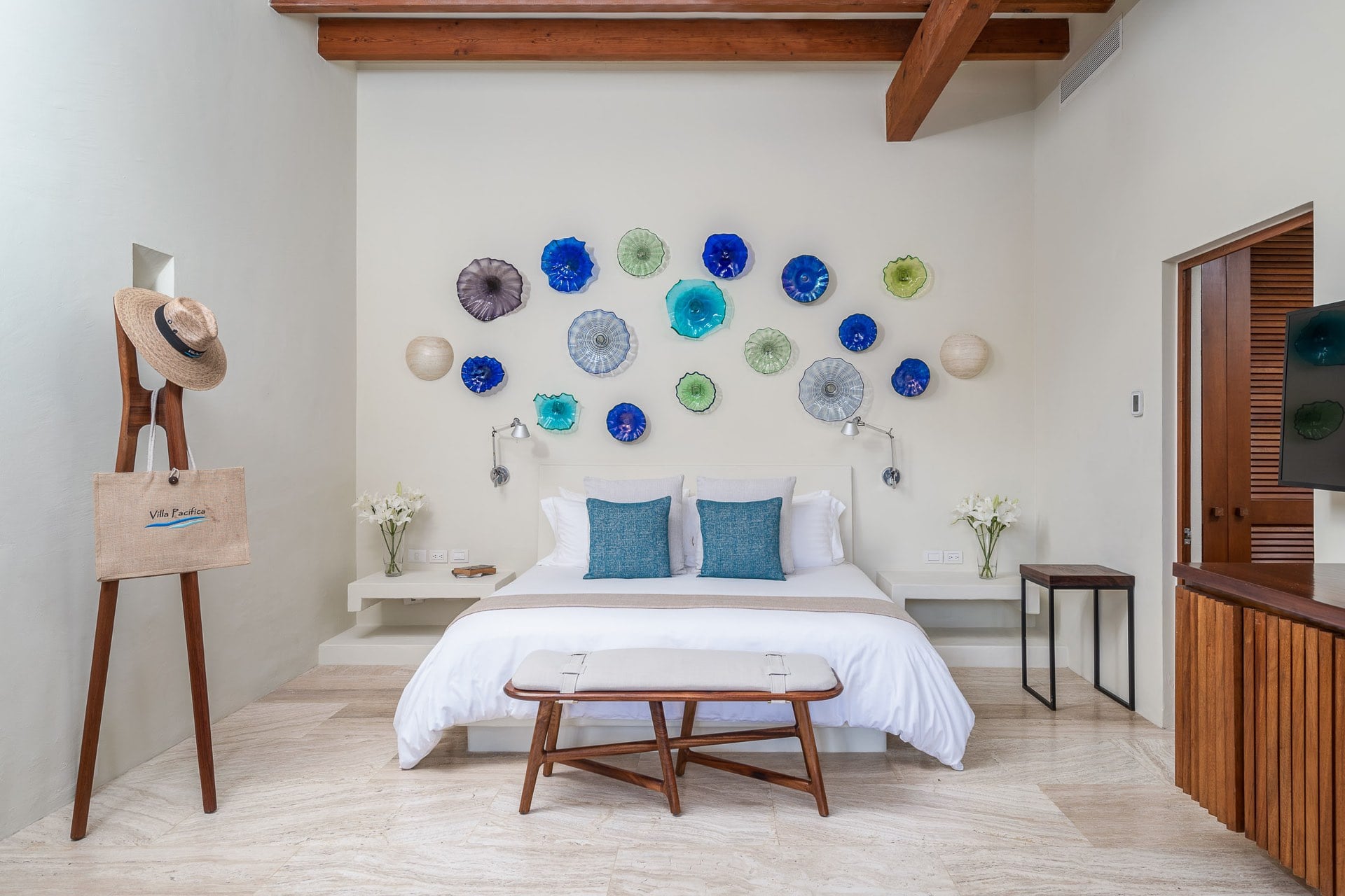 Guest suite king bed beneath a gallery of blown-glass disc wall art in cobalt and green, wood-beam ceiling, teak bench at foot, Villa Pacífica branded tote on coat rack, Villa Pacífica, Ranchos Estates, Punta Mita.