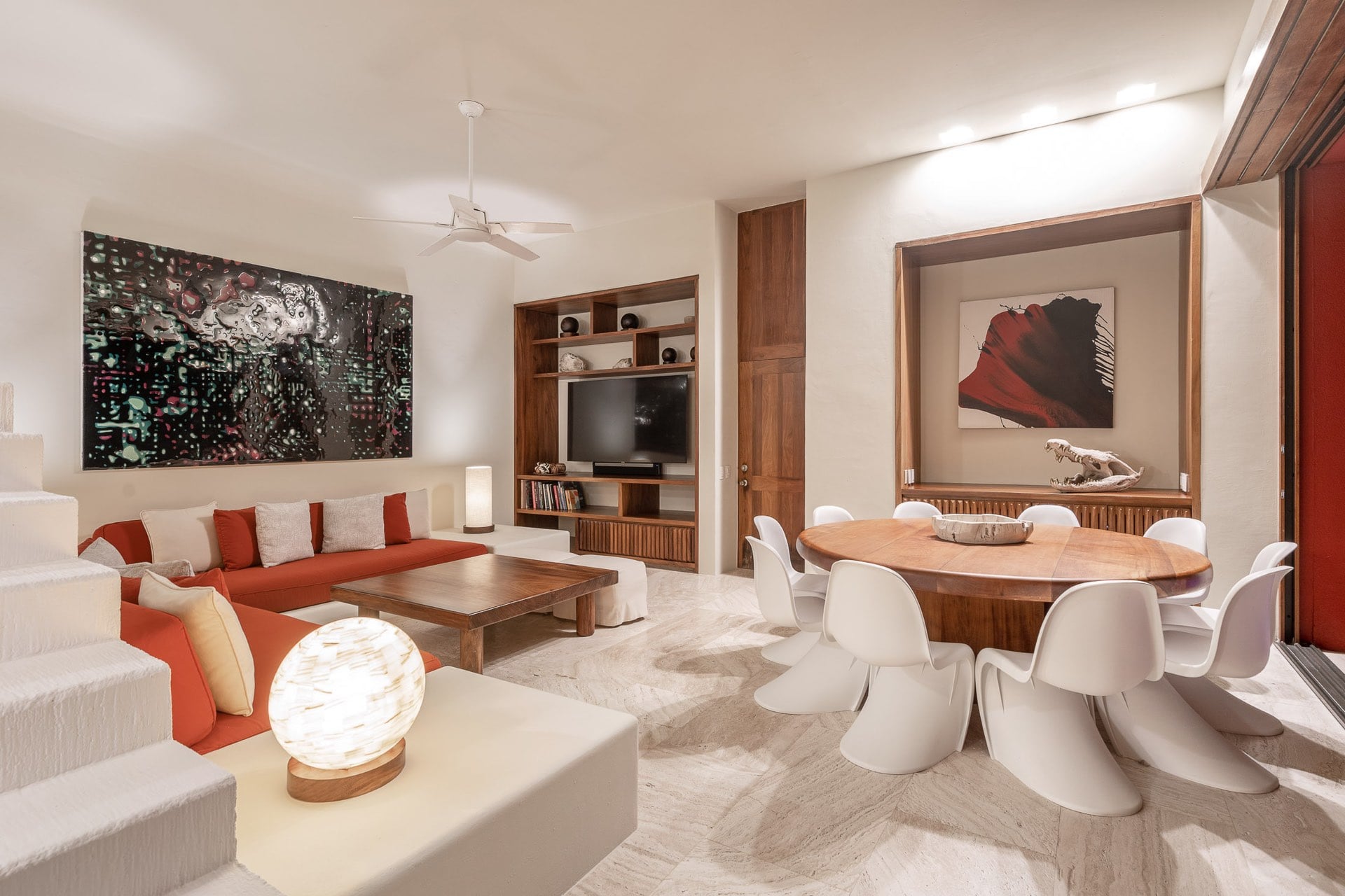 Open living area with terracotta sectional sofa and walnut coffee table adjoining a round walnut dining table with eight white Panton chairs, built-in media shelving and bold abstract canvas on wall, Villa Pacífica, Ranchos Estates, Punta Mita.