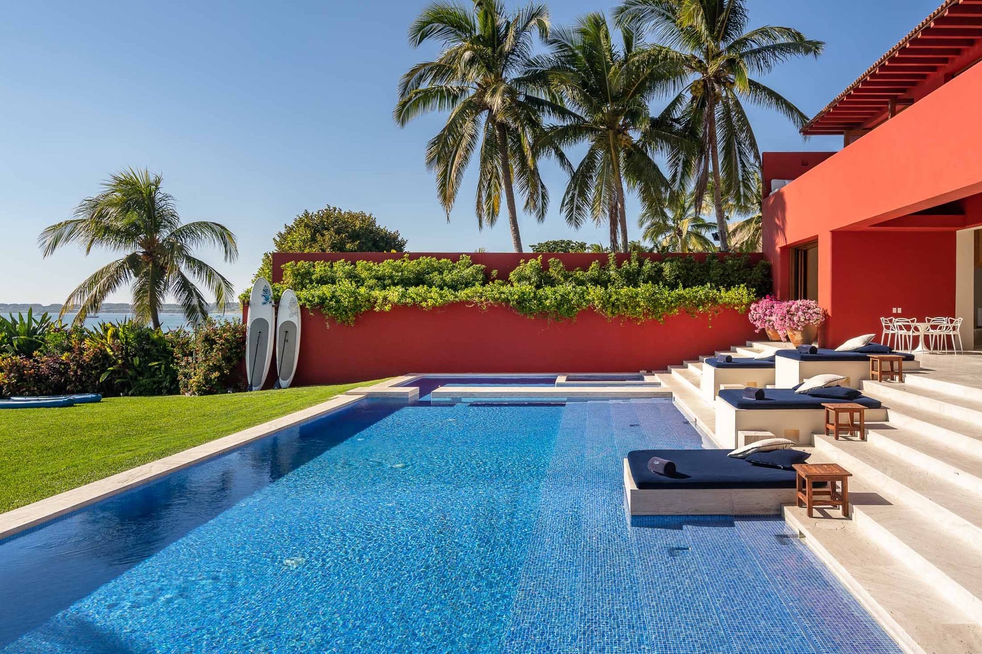 Rectangular mosaic pool with adjacent hot tub, stepped terrace with navy sun loungers, two paddleboards against coral-red boundary wall, and ocean at left, Villa Pacífica, Ranchos Estates, Punta Mita.