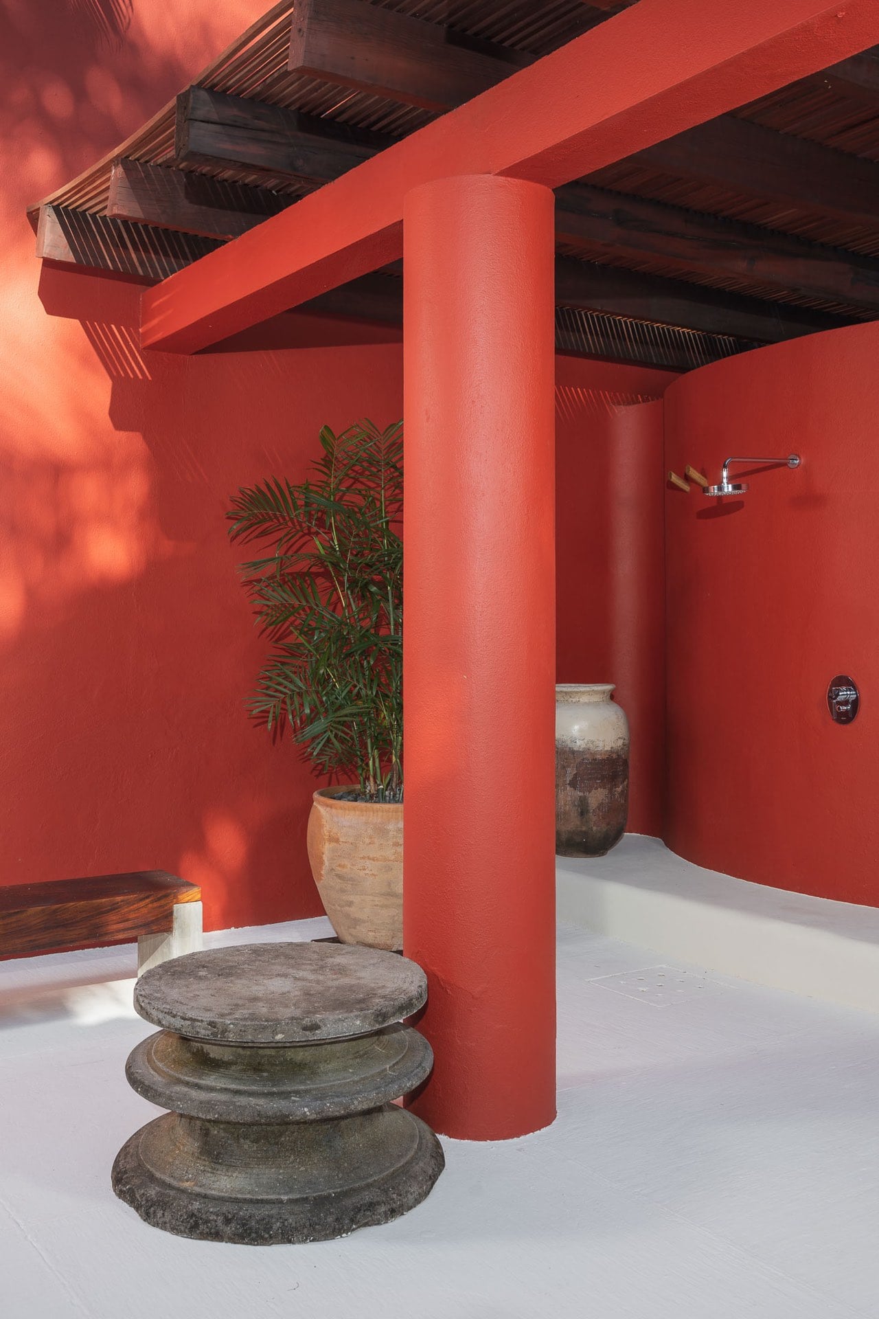 Outdoor shower alcove within coral-red stucco walls and slatted wood pergola, rain showerhead on wall, antique clay urns, potted palm, and carved stone pedestal in foreground, Villa Pacífica, Ranchos Estates, Punta Mita.